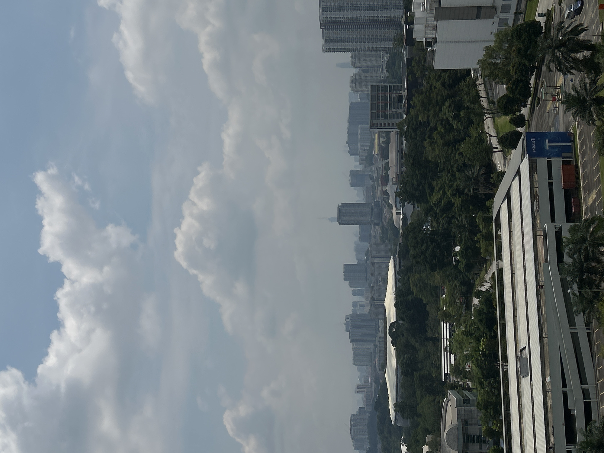 The image presents an elevated view of Kuala Lumpur, Malaysia, under a cloudy sky. The city skyline is composed of numerous high-rise buildings. Vegetation including trees and palms are scattered throughout the urban landscape. A modern white building with a curved facade and linear design is visible in the foreground. Roads and parked vehicles are also present. A sign with the word "MRANTI" is partially visible.