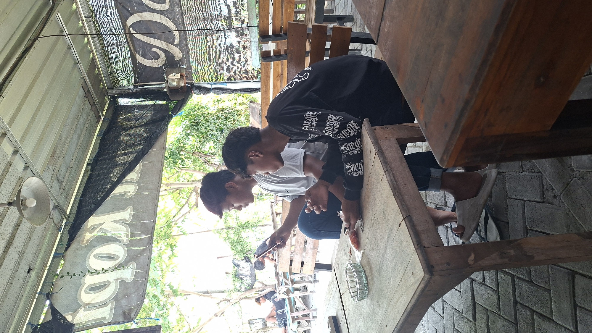 Two young men are seated at a wooden table under a covered outdoor seating area, likely a warung or cafe, in Ngimbang, Indonesia. Both men are looking at their mobile phones, engrossed in their devices. The man on the left is wearing a light grey t-shirt and blue jeans, while the man on the right is wearing a black long-sleeved shirt with white lettering.
On the table between them, there is a glass ashtray, a small packet, and what appears to be a piece of food, possibly a fish. The table surface is worn and weathered. Behind them, wooden benches and tables are visible, some occupied by other patrons in the background who are also engaged with their phones.
The environment suggests a casual, open-air setting. Lush green trees and foliage are visible beyond the covered area, indicating an outdoor location. A large banner with Indonesian text, possibly advertising coffee ("Kopi"), is partially visible hanging above the seating area, along with a corrugated metal roof and a light fixture. Another banner with the word "Da" is visible on the right. The ground is paved with interlocking bricks. The overall scene depicts a relaxed atmosphere where people are spending time together while utilizing their technology.