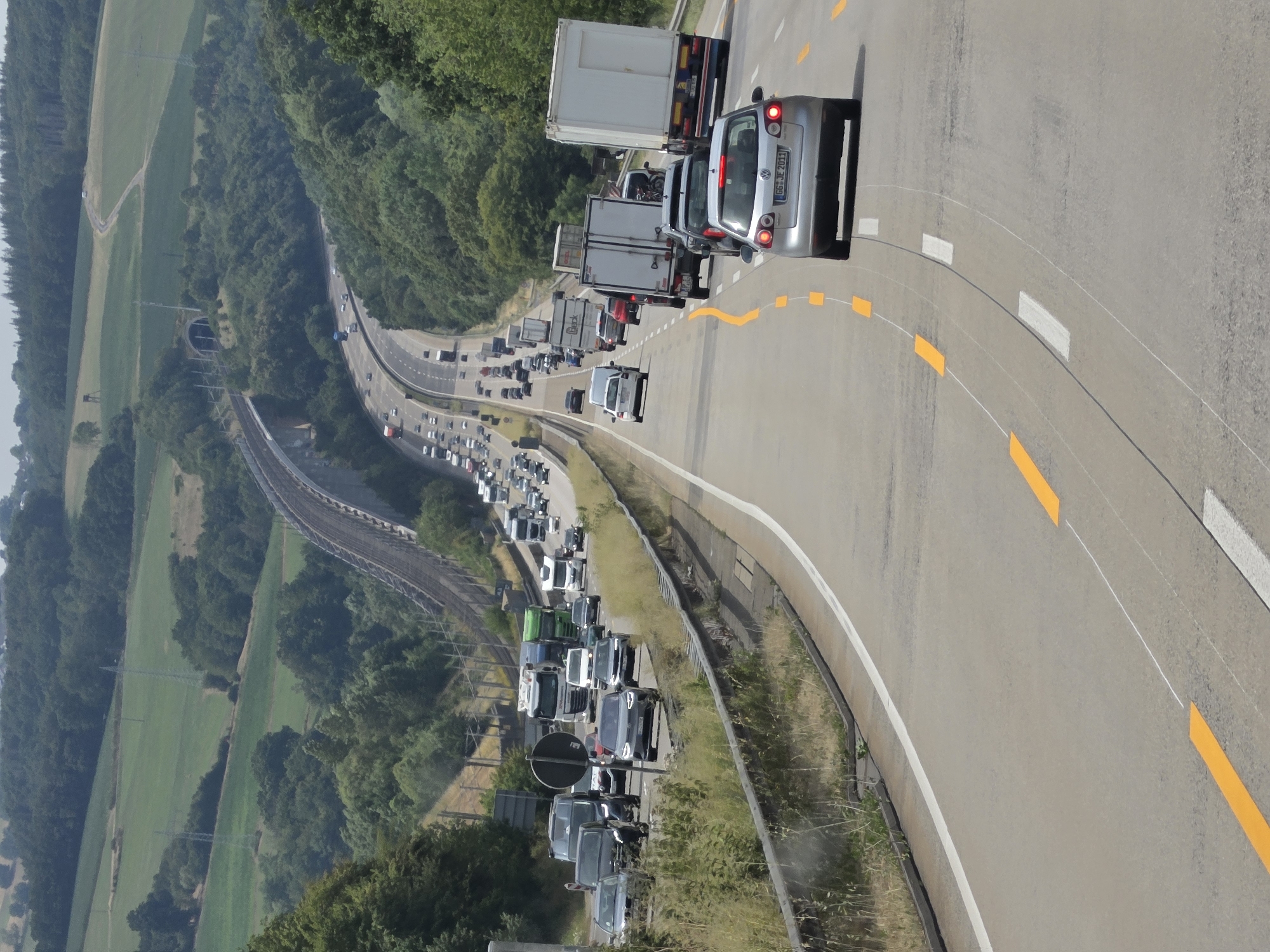 The image shows a highway in Neustadt (Wied), Germany, with moderate traffic congestion. Several cars and trucks are visible on the road, with some vehicles appearing to be stationary or moving slowly. The highway is surrounded by lush green hills and forests, characteristic of the German countryside. A bridge and a tunnel are also visible in the distance, suggesting a section of the Autobahn. The weather appears to be clear and sunny, with a few clouds in the sky. The overall scene depicts a typical summer day on a German highway.