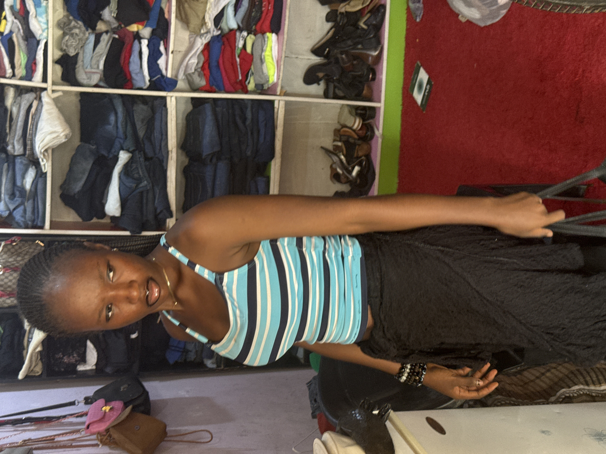The image shows a woman in a striped tank top and dark skirt in front of a closet. The closet is filled with neatly folded clothes and shoes. The floor is covered in a red rug. There are handbags and purses on a lower shelf. The location is in Abuja, Nigeria.