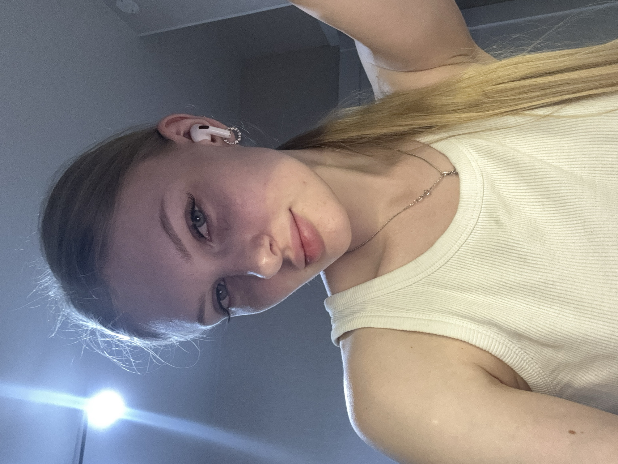 A close-up, slightly rotated selfie photograph features a young woman with fair skin and blonde hair pulled back in a ponytail. She is wearing a white ribbed tank top and a delicate silver necklace. An Apple AirPods Pro is visible in her left ear, with a dangling jeweled charm attached. The background is a muted, grey-toned wall, suggesting an indoor setting. A bright light source is visible towards the lower left of the frame.
While the image provides details about the woman and her immediate surroundings, there is no information within the image to determine the location or any indication of its relation to Perm, Russia. Without additional context, it is impossible to place the photograph in that city.