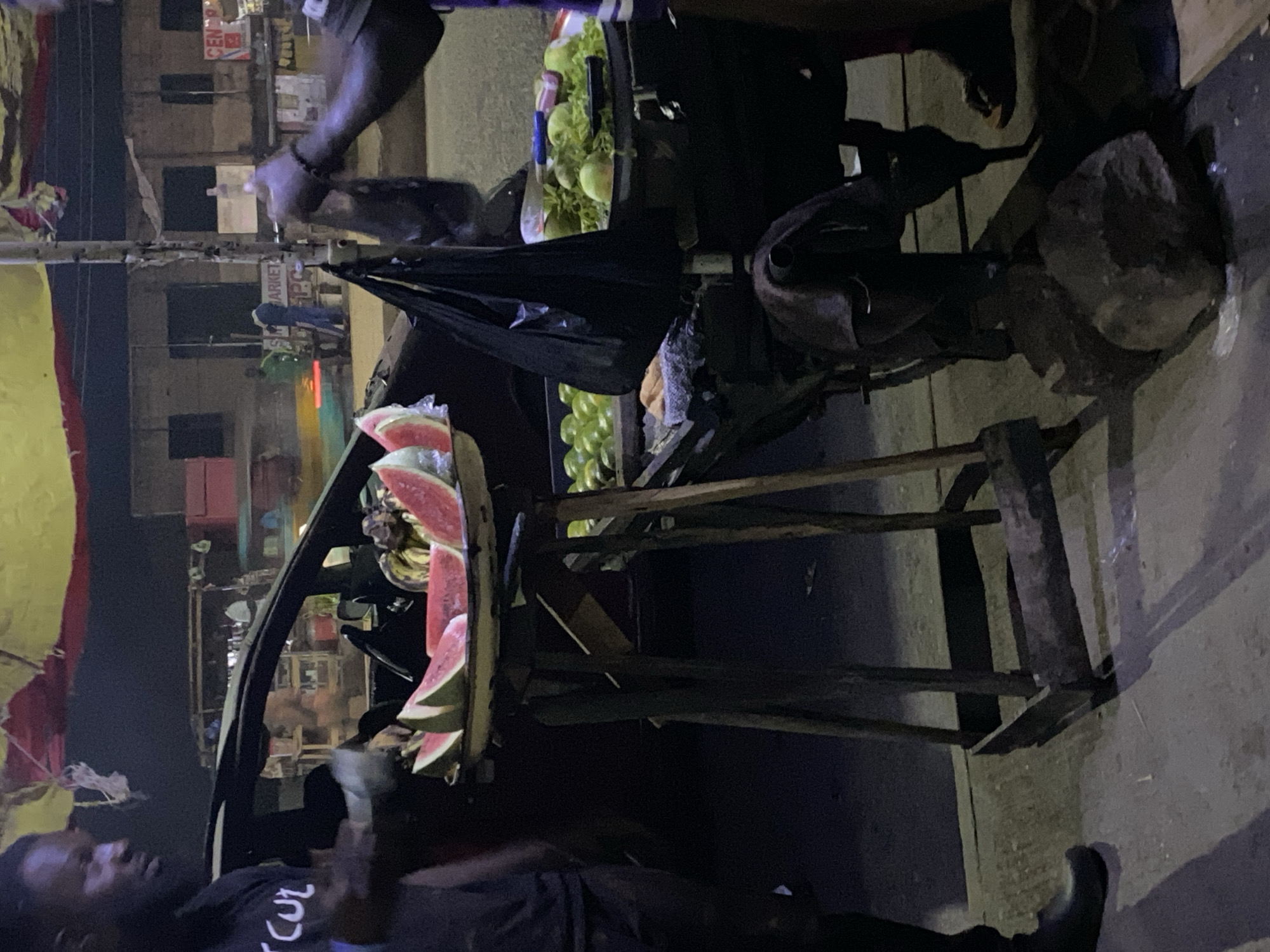 This low-light photograph, likely taken at night in Bukuru, Nigeria, captures a vibrant street vending scene. The central subject is a wooden fruit stand laden with fresh produce. Visible on the stand are numerous slices of red watermelon, along with bunches of green grapes and other green fruits, possibly apples or mangoes. Dark plastic bags hang from the stand's structure.
Two individuals are primarily visible. In the foreground, on the left, a man wearing a dark t-shirt with partial white text (possibly "CUT") is seen, his face dimly lit. His right hand appears to be holding something. Another person's arm and hand are visible near the fruit stand in the upper middle, suggesting they are handling the produce or serving a customer.
The background features blurred street activity, including other figures, one appearing in blue and another in green, alongside buildings and structures typical of an urban environment. Artificial lighting from shops or streetlights illuminates parts of the scene. A partial car is visible on the left side, behind the man in the foreground.
Notable textual details include the word "MARKET" clearly visible on a sign in the background, with "PPC" below it, and "CENDE..." on another distant sign. The overall ambiance suggests a busy market or roadside stall operating during evening hours.