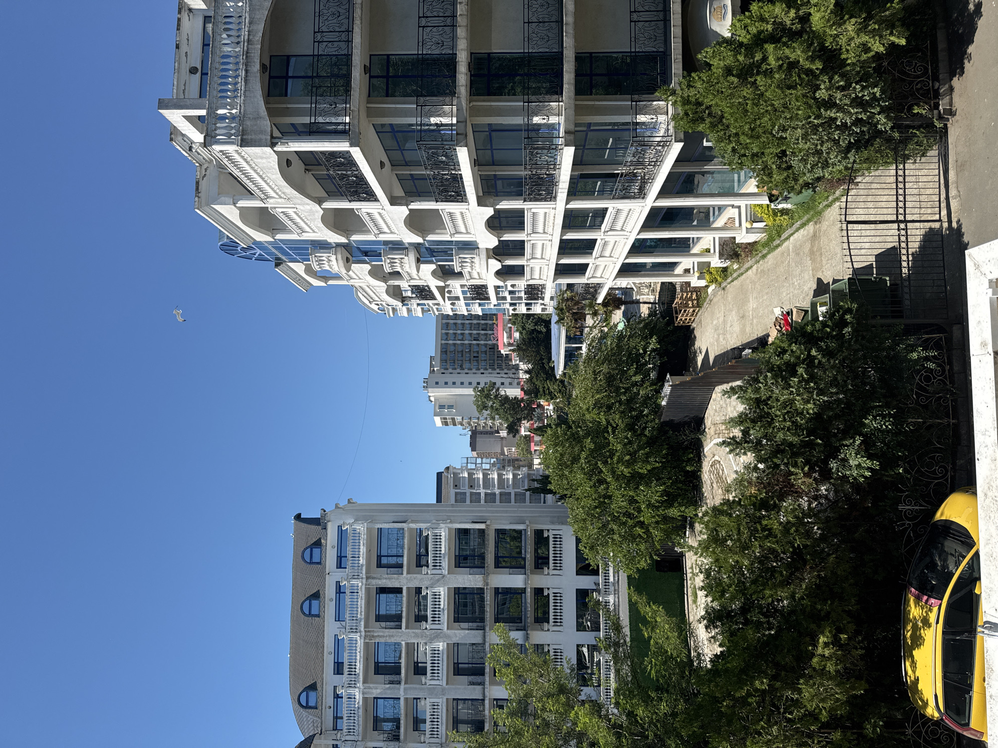 This image depicts an urban scene on a clear, sunny day. In the foreground, a vibrant yellow car is parked, partially obscured by lush green trees and bushes. A black metal fence runs along the edge of the property.
Dominating the midground and background are several multi-story buildings with white facades, balconies, and numerous windows. The architecture appears modern, with some buildings featuring curved elements and ornate railings. The building on the right is closer and larger, showcasing multiple levels of balconies with intricate black metal railings and large, blue-tinted windows. To its left, another white building of similar style is visible, featuring arched windows and a mansard roof with dormer windows. In the distance, taller, more uniform buildings rise against the blue sky.
A paved pathway leads towards the buildings from a gate in the foreground. Greenery, including trees and manicured bushes, is interspersed throughout the scene, suggesting a well-maintained urban environment, possibly a resort or residential area. A single bird is visible in flight against the clear blue sky. The overall impression is one of a bright, warm day in a developed, possibly coastal, urban setting.