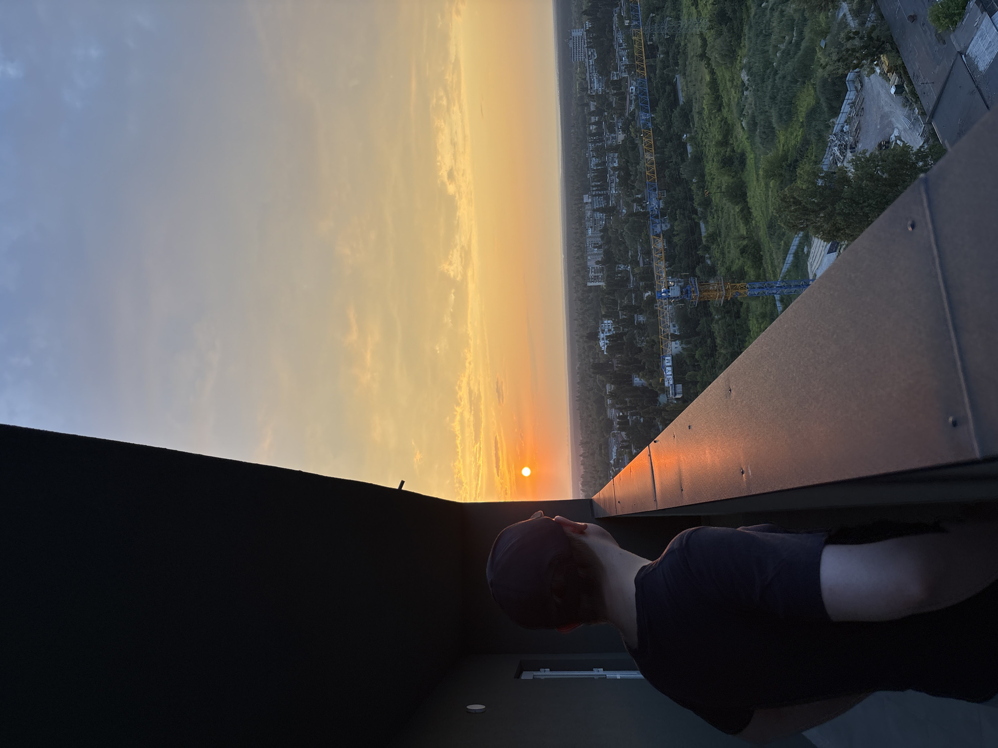 A young person wearing a dark t-shirt and a baseball cap stands on a balcony overlooking a city at sunset. The person is facing away from the camera, looking out at the horizon where the sun is setting in a vibrant orange and yellow glow. The sky is filled with soft clouds. Below the balcony, a sprawling cityscape is visible, with many trees and buildings. A construction crane is prominent in the middle ground, suggesting ongoing development in the area. The balcony itself has a dark, modern railing and a dark wall to the left, casting a shadow over part of the scene. The lighting suggests it is late afternoon or early evening. The scene is captured from a high vantage point, offering a panoramic view of the urban landscape.