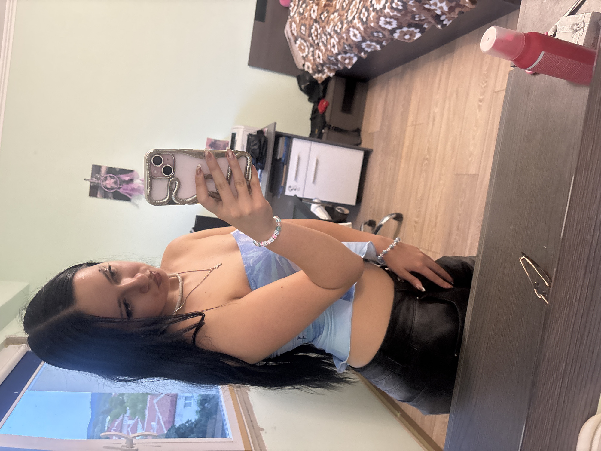 A young woman with long, dark hair and fair skin takes a mirror selfie in a bedroom. She is wearing a light blue, strapless crop top and black, low-waisted pants. She has on a necklace and bracelets. She is holding a pink-cased smartphone in her right hand, which is angled to capture her reflection. To her left, a window offers a view of houses and hills under a cloudy sky. In the background, a bed with a patterned comforter, a desk with a printer, and a file cabinet are visible. The room appears to be a domestic setting.