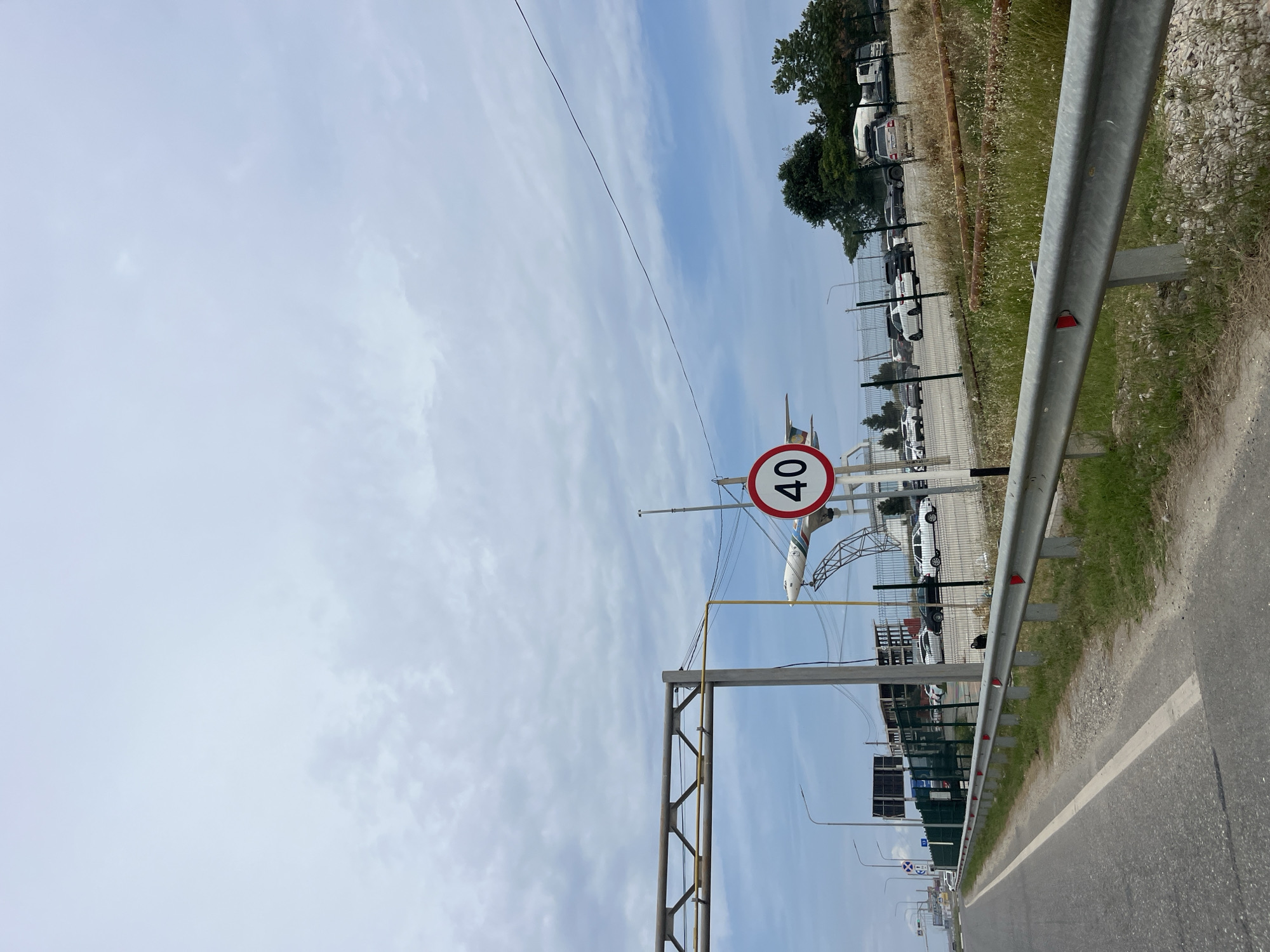 The image shows a road with a metal guardrail on the right side. Beyond the guardrail, there is a fenced-off area with several cars parked. In the background, a model airplane is visible mounted on a structure, and what appears to be construction or an unfinished building is present. A speed limit sign indicating "40" is also visible. The sky is cloudy. The scene suggests an area near an airport or a transportation hub, potentially in the city of Tural-Sul'fat, Russia.