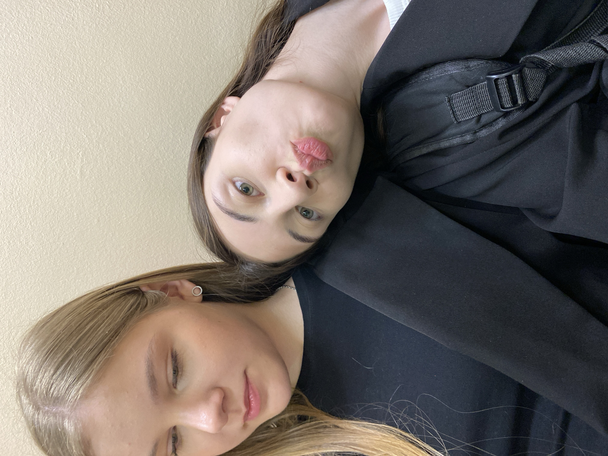 Two women are posing for a selfie. The woman on the top has long, brown hair and is wearing a black blazer and what appears to be a backpack. She is making a kissing face. The woman on the bottom has long, blonde hair and is slightly smiling. Both women are fair-skinned. The background is a light beige wall. There is no indication of the city of Votkinsk, Russia.