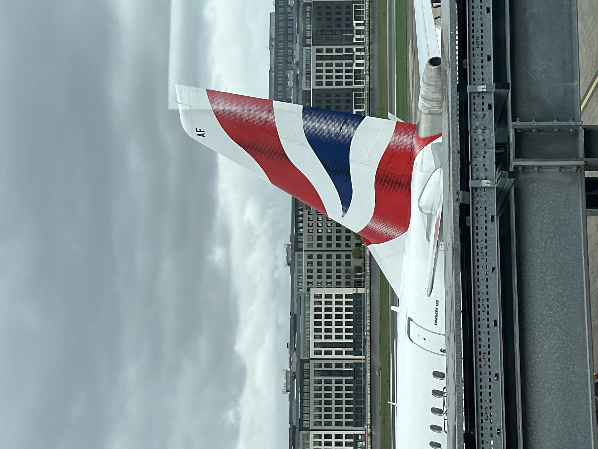 The image shows the tail of a British Airways Embraer 190 aircraft, visible through a window. The tail features the Union Jack flag design, with the registration "AF" faintly visible. In the background, a row of modern apartment buildings is observed under a cloudy sky. The photograph was taken from within Heathrow Airport in London, United Kingdom, with a metal structure, likely part of the airport's infrastructure, in the foreground.