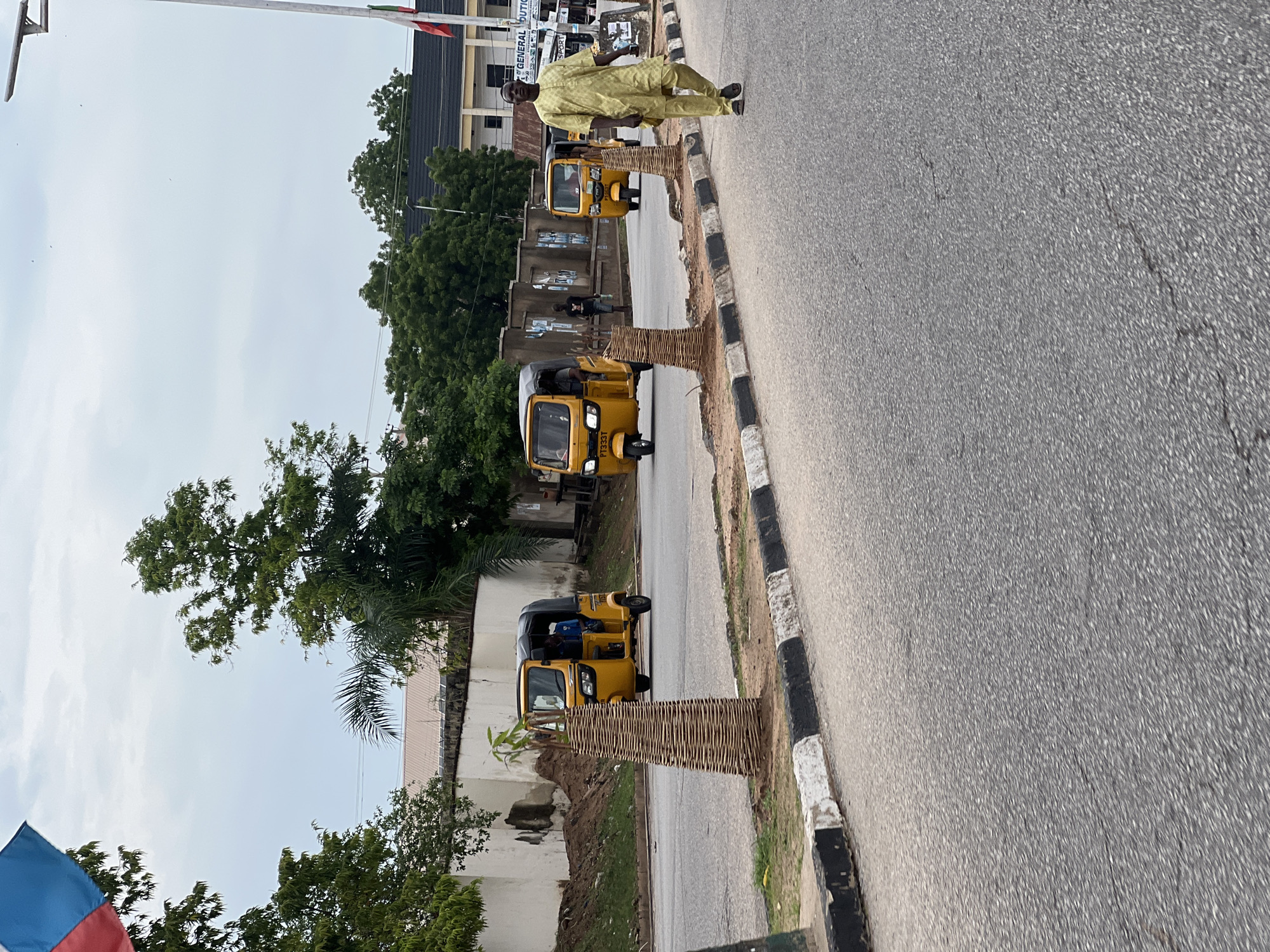 In Jalingo, Nigeria, on a paved road with a black and white striped curb, a man in a pale yellow traditional garment walks away from the camera. Behind him, three yellow auto-rickshaws, also known as Keke Napep, are visible. The first auto-rickshaw is closer to the camera, with a driver and one passenger inside, turning left. The second auto-rickshaw is further down the road, also with a driver visible. The third auto-rickshaw is even further down the road and its occupants are not clearly discernible. The background features a white building with a corrugated roof, trees, and a stone wall. A flag with red, blue, and white colors is partially visible in the upper left corner of the image. The sky is overcast.