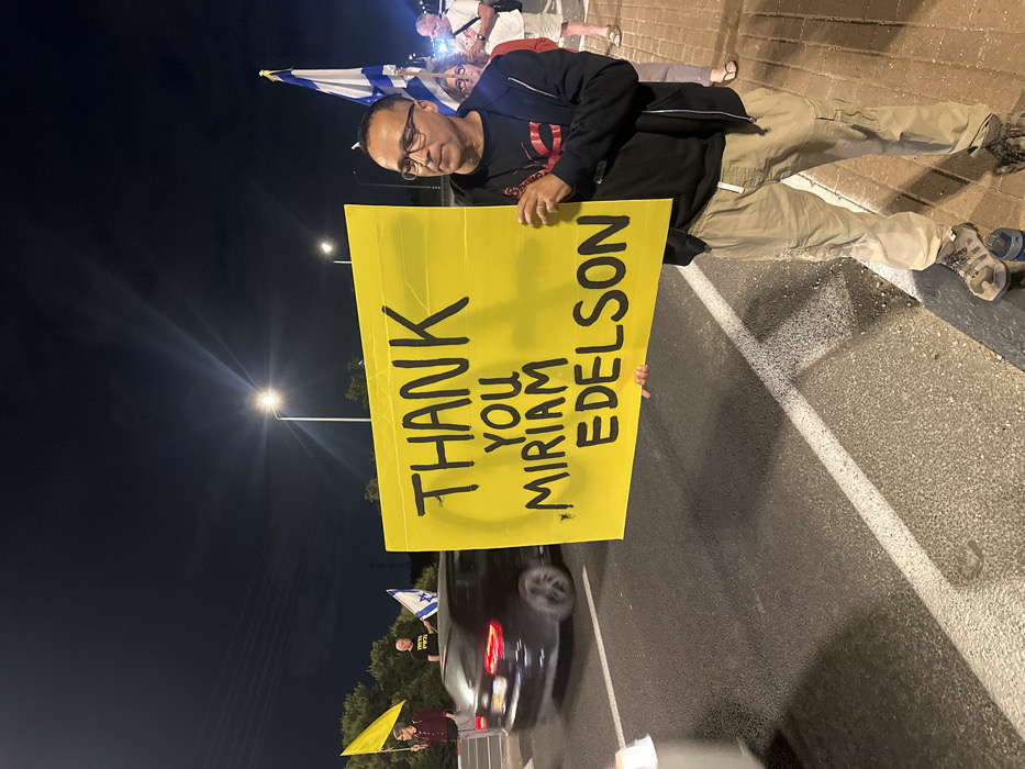 A nighttime image taken in Mishmar Hasharon, Israel, captures a roadside gathering. In the foreground right, a man, appearing to be in his 40s or 50s, with glasses, a dark hooded jacket, and light cargo pants, holds a large, bright yellow sign horizontally. The sign displays "THANK YOU MIRIAM EDELSON" in bold, black, hand-drawn capital letters. To the man's right and behind him, partially visible, are at least two other individuals, one of whom holds an Israeli flag. Across the two-lane asphalt road to the left, two additional individuals are visible; one holds a solid yellow flag, and the other holds an Israeli flag. A dark-colored sedan is captured mid-motion on the road, indicating active traffic. The environment is illuminated by multiple overhead streetlights, casting light on the participants and the road, which features white lane markings. The sky above is dark and clear, with distant foliage visible on the left.