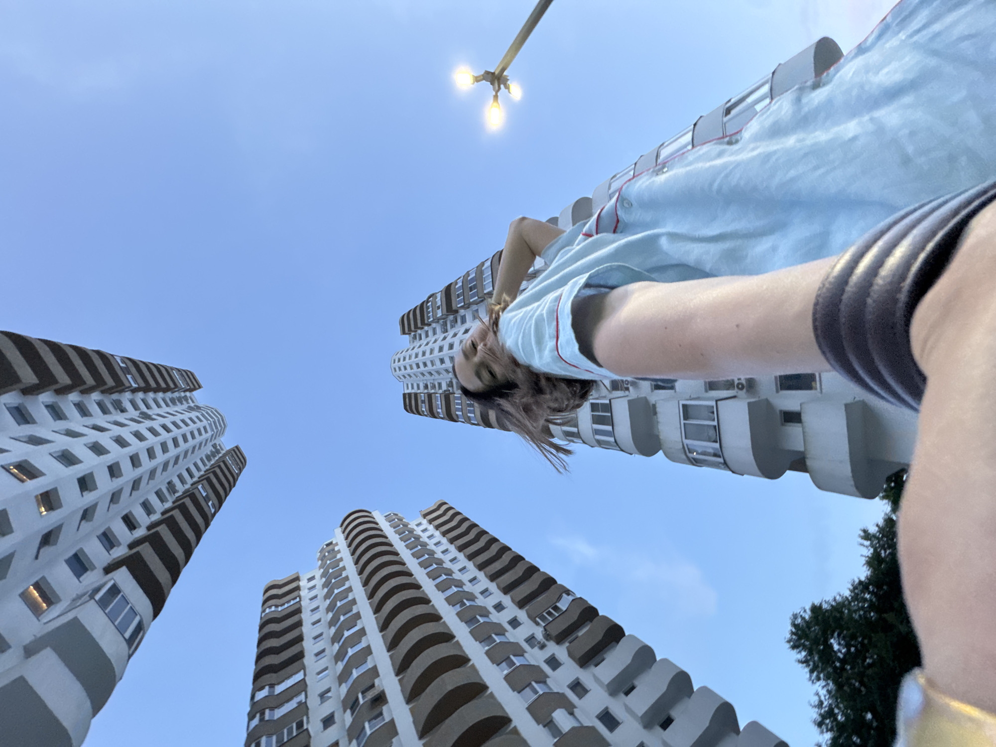 A low-angle, wide-shot photograph taken from below shows three tall, modern apartment buildings against a clear blue sky. The buildings are characterized by their curved balconies and numerous windows, with a light-colored facade and dark-colored decorative bands. In the foreground, a person is captured in a selfie-style shot, with their arm extending towards the camera. The individual, appearing to be female, is wearing a light blue garment with red trim and has their hair flowing. They are looking towards the camera with a slight smile and are positioned in front of the tallest of the three buildings. To the right, a street lamp with three illuminated bulbs is visible. Trees are also present in the lower left corner of the frame. The overall setting suggests an urban environment, possibly in Sochi, Russia, given the architectural style of the buildings and the bright, clear sky indicative of a temperate climate.