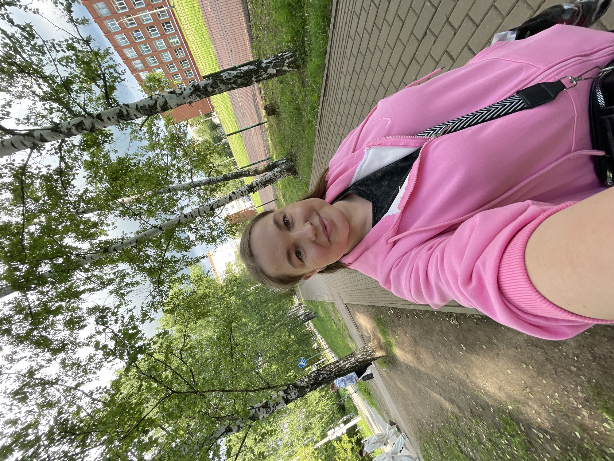 A selfie taken by a young woman outdoors in Izhevsk, Russia, is presented. The woman occupies the lower-middle portion of the frame, looking directly at the camera with a neutral to slightly content expression. She is wearing a vibrant pink zip-up hooded sweatshirt over a dark grey or black top, and a black and white striped bag strap is visible across her torso.
The setting is an outdoor green space, likely a park or common area. A paved path, made of light-colored bricks, runs parallel to the woman and is lined by several birch trees featuring white bark and abundant green leaves. Beyond the path lies a grassy expanse. In the background, a multi-story building made of red brick with numerous windows stands prominently. To the right of the building, a green fence is visible, suggesting an athletic field or enclosed area. A blue road sign with a white arrow is partially seen on a pole near a tree.
The lighting indicates it is daytime, with an overcast or partly cloudy sky providing soft, diffused illumination. The overall scene conveys a casual and relaxed atmosphere. No discernible text is visible on any objects within the image.