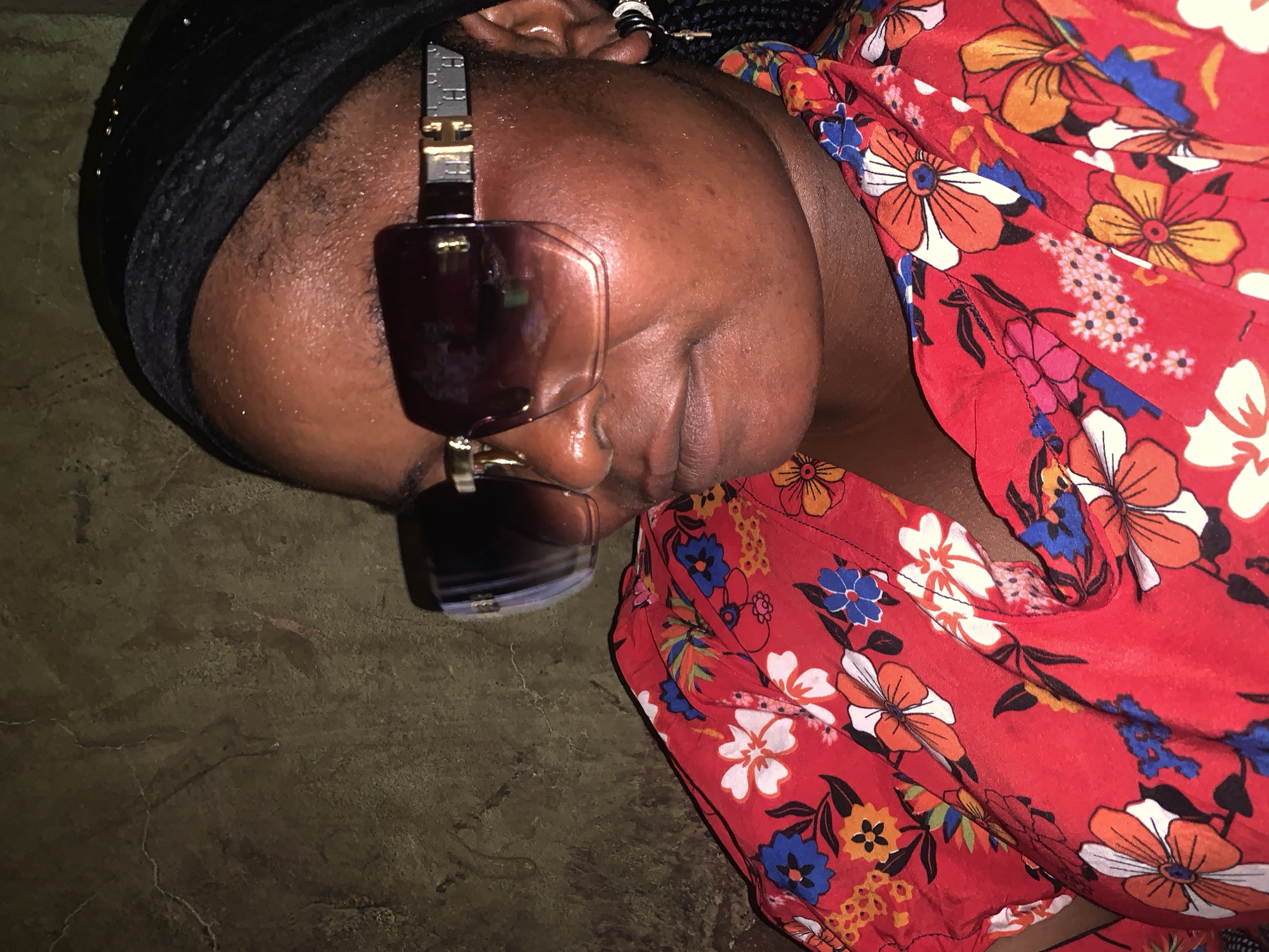 The image shows a close-up, overhead view of a person lying down. The person is wearing a black headscarf and large, dark sunglasses with a metallic accent on the temples. They are also wearing a red dress with a vibrant floral pattern featuring white, orange, and blue flowers. The person's face is partially obscured by the sunglasses and their head is tilted. Their skin appears to be dark, and there is visible sweat on their forehead. The background is a textured, dark surface, possibly concrete or earth. The lighting suggests it may be indoors or during the evening, as there are shadows. The overall impression is one of rest or relaxation. There is no visible text in the image. The provided location context is Boi A, Nigeria.