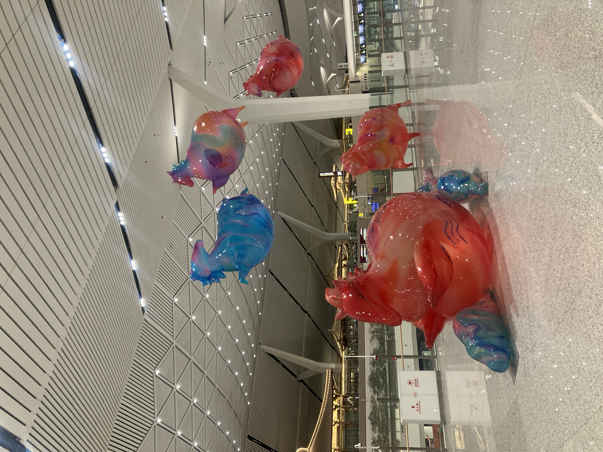 The image shows a modern airport interior with several colorful, abstract, animal-like sculptures suspended from the ceiling. The sculptures are glossy and appear to be made of plastic or resin. They are various colors including red, blue, purple, and orange. The airport features a high ceiling with a grid-like design. Large windows line the walls, reflecting light onto the polished floor. The image appears to be taken at the Xianyang International Airport in the Weicheng District of Xianyang, China, as confirmed by the presence of these sculptures within the airport.