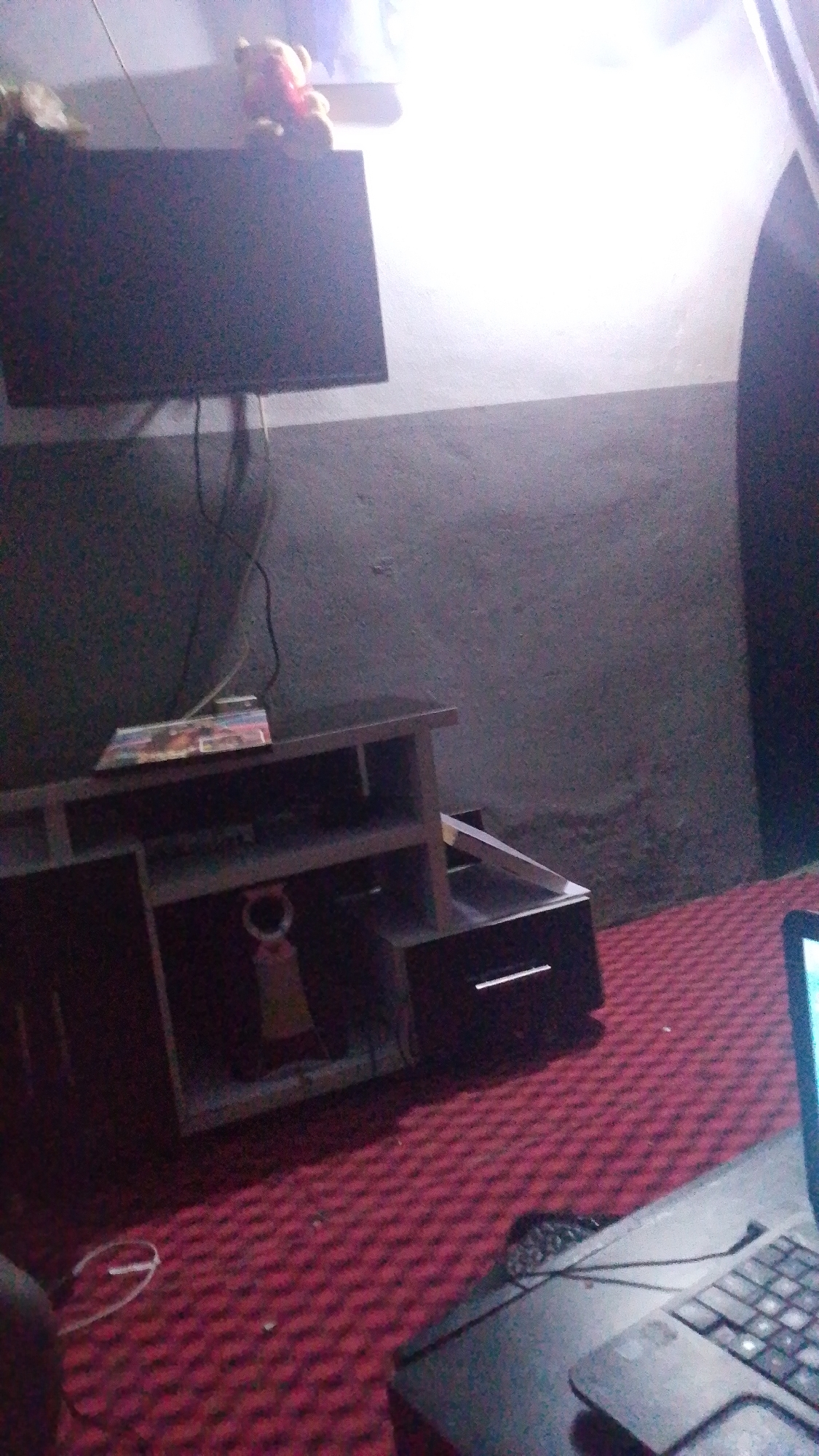 In a dimly lit room, a flat-screen television is mounted on the wall above a dark wooden media console. The console has multiple shelves and drawers, with a few items placed on the top shelf, including what appears to be a book or case with colorful graphics and a small, light-colored teddy bear is sitting on top of the television. The room is carpeted with a red, patterned rug, and a laptop is open on the floor to the right of the image. The wall behind the television is a dark, textured surface, and the lighting in the room appears to be coming from a bright light source to the right, creating a strong contrast and casting shadows.