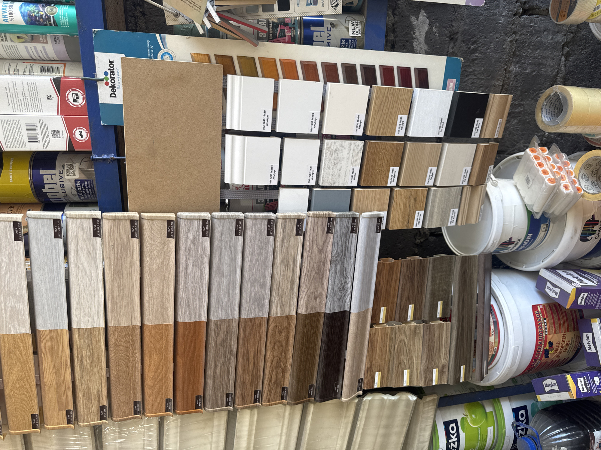 The image shows a retail display of flooring and building materials in a store. The primary focus is on stacked planks of laminate flooring with various wood grain patterns and colors, organized in horizontal rows. Some planks are half wood grain and half a solid color, likely representing different styles or finishes.
To the right of the flooring display, there are samples of skirting boards, also in different finishes and colors, some with labels containing product information. In the background, shelves are stocked with paint cans, adhesives, and other building supplies. Notably, there are cans of "Nobel" paint, and a can labeled "AKVARYUM %100 SILIKON". A prominent sign for "Dekorator" is also visible.
In the foreground and to the left, there are large buckets of paint or other building materials, along with smaller boxes labeled "Metylan" and a bottle of dark liquid. Rolls of tape are also present in the lower right portion of the image.
The setting appears to be an indoor retail environment, possibly a hardware store or a specialized flooring shop. The presence of Russian and possibly Georgian text on some of the product labels suggests the location might be in a region where these languages are common. Given the request to include the city of Tbilisi, Georgia, this scene is depicted as occurring within a retail establishment in Tbilisi. The image is static and does not show any people actively engaged in transactions, but rather the products themselves are the subjects of the photograph.