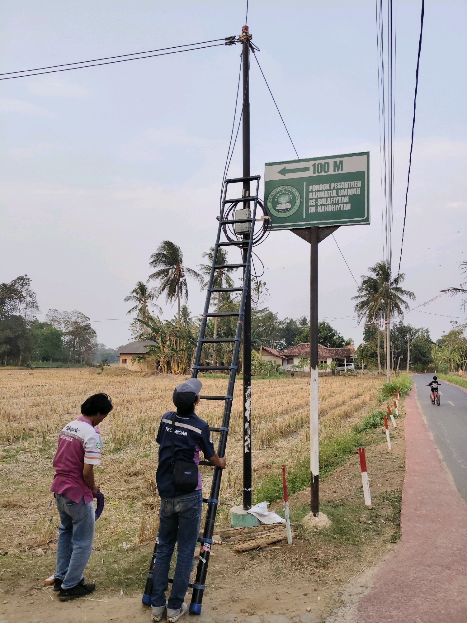 In the city of Sidosari, Indonesia, an outdoor scene unfolds with a power pole at its center, equipped with a ladder and various electrical components. Two individuals, likely technicians, are present near the pole. One, wearing a pink and white shirt with "TECHNICIAN" visible on it, stands with a purple coil of wire. The other, in a blue shirt also marked "TECHNICIAN" and a grey cap, stands on the ladder, appearing to be working on the pole's equipment. A green directional sign with "100 M" and text in Arabic script is mounted on the pole, indicating the direction towards "Pondok Pesantren Rahmatul Ummah As-Salafiyyah An-Nahdhiyyah."
The environment surrounding the pole is a rural landscape. A dry, harvested rice field stretches behind the individuals. In the background, a small building with a tiled roof is visible, along with scattered trees, including palm trees. A paved road runs alongside the field, and a young person on a red bicycle is seen riding away from the camera on this road. The sky is overcast with a pale blue hue. Several small red and white markers are placed along the edge of the road.