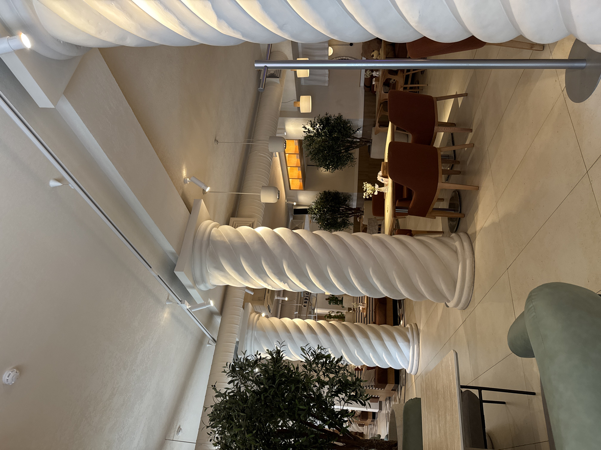 The image depicts the interior of a restaurant, likely Capital, located in Almaty, Kazakhstan. The scene is dominated by two large, white, twisted columns that occupy the foreground and midground. In the background, various seating arrangements are visible, including tables with chairs and booths, some with patrons. Several artificial olive trees are placed strategically throughout the restaurant, adding a touch of greenery. The lighting appears to be a mix of ambient and spotlight illumination, creating a warm and inviting atmosphere. The overall aesthetic suggests a modern and chic dining establishment.