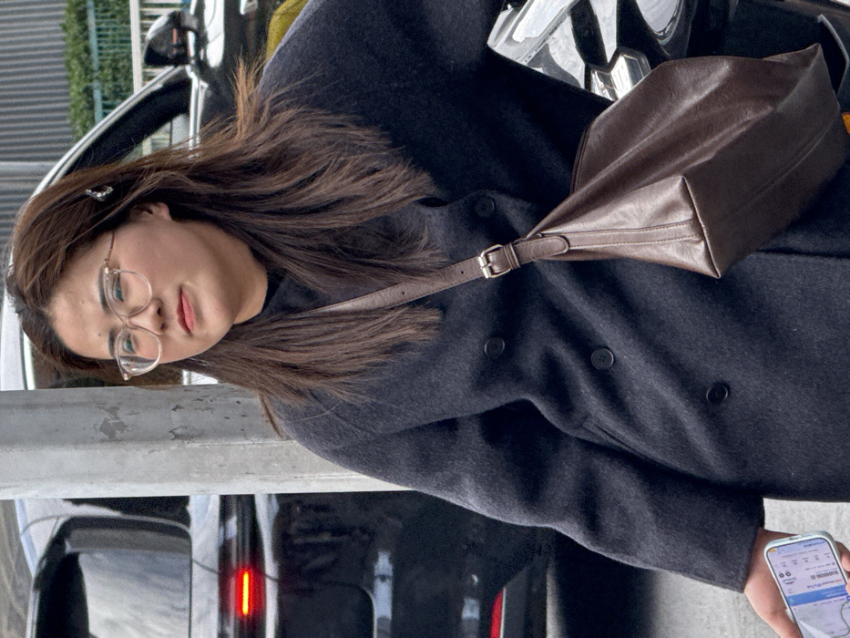 A woman with glasses and a hair clip is standing outside, holding a smartphone in her hand. She's wearing a dark, double-breasted wool coat and has a brown leather shoulder bag slung across her body. A black vehicle with illuminated tail lights is visible behind her. The woman appears to be looking down at her phone.