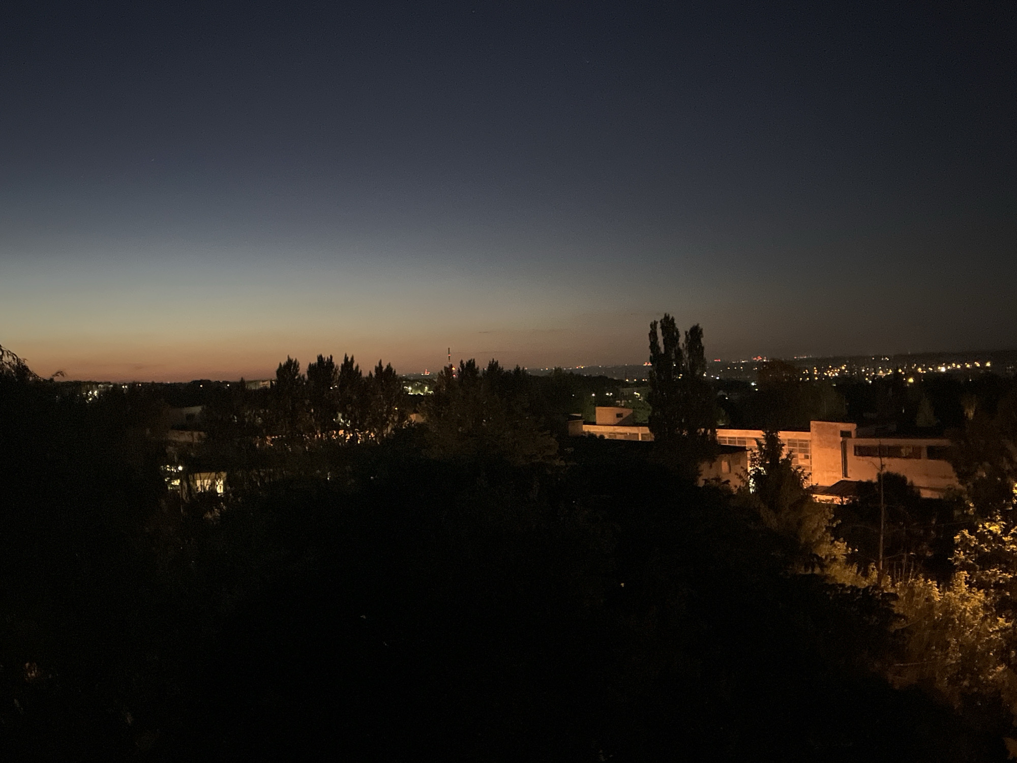 Here's a description of the image:
The image depicts a nighttime cityscape. The sky transitions from a dark blue overhead to a lighter orange glow near the horizon. Silhouetted trees line the midground. In the distance, there are buildings and scattered lights, suggesting a city. The foreground is very dark, with indistinct shapes possibly representing vegetation. A few buildings are visible, illuminated by interior lights. The image does not offer any specific identifiers to definitively locate it within a city of Unknown, Unknown.