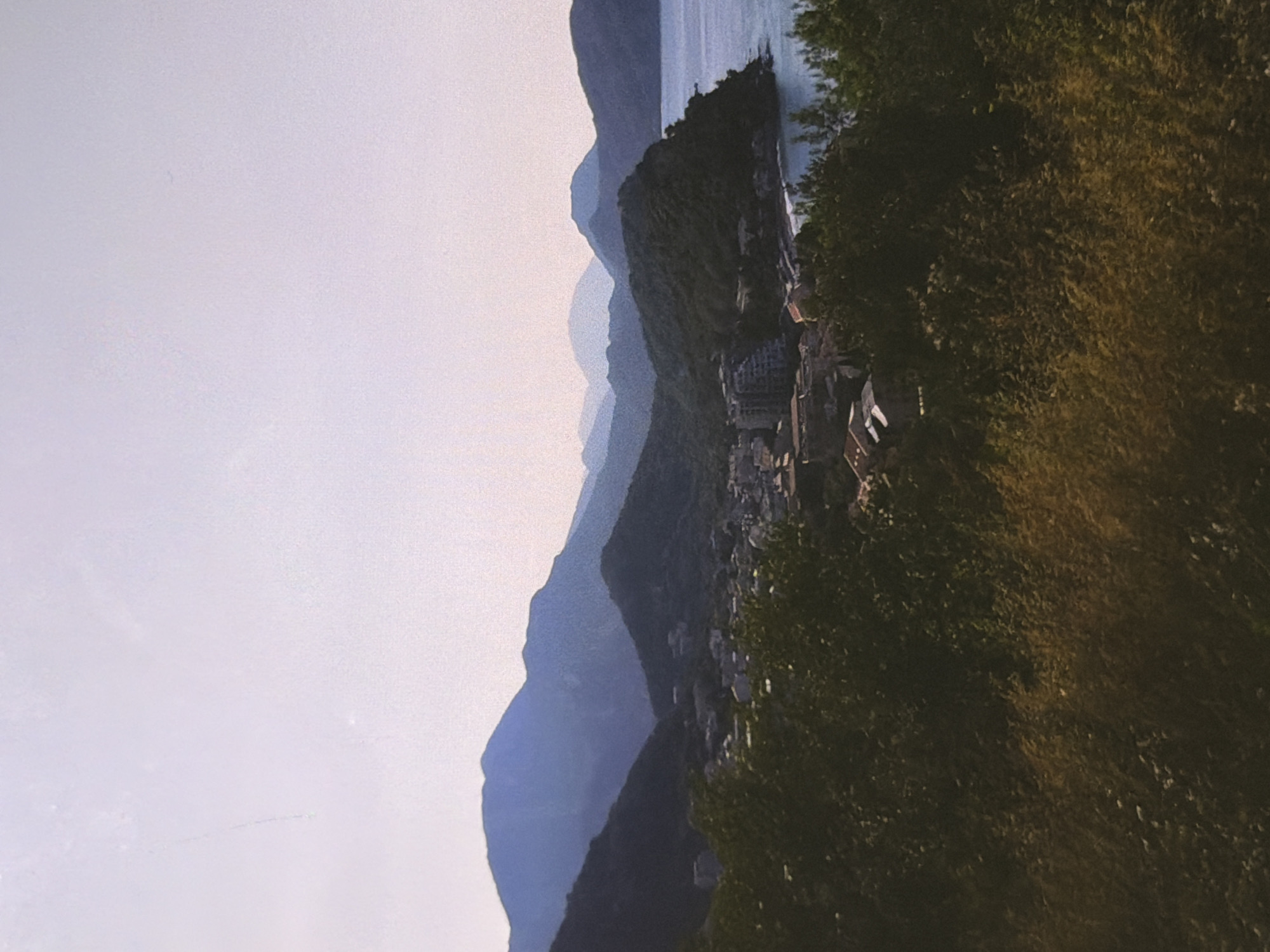 A panoramic view captures a coastal town nestled between lush green hills and the shimmering sea. The sky above is a pale, washed-out blue, hinting at either dawn or dusk. In the foreground, dense foliage with dry, golden grass obscures the lower portion of the frame. Beyond this, a cluster of buildings, appearing to be houses and perhaps a hotel, are spread across a slope leading down to a small bay. The structures have terracotta roofs and white walls. The mid-ground is dominated by rolling, dark green hills, some of which are bare, while others are thickly forested. A series of progressively distant mountain ranges form the backdrop, their blue hues deepening with their recession into the horizon. The sea, a tranquil expanse of blue, meets the coastline and extends to the right of the frame. The scene is still and serene, with no visible human activity.