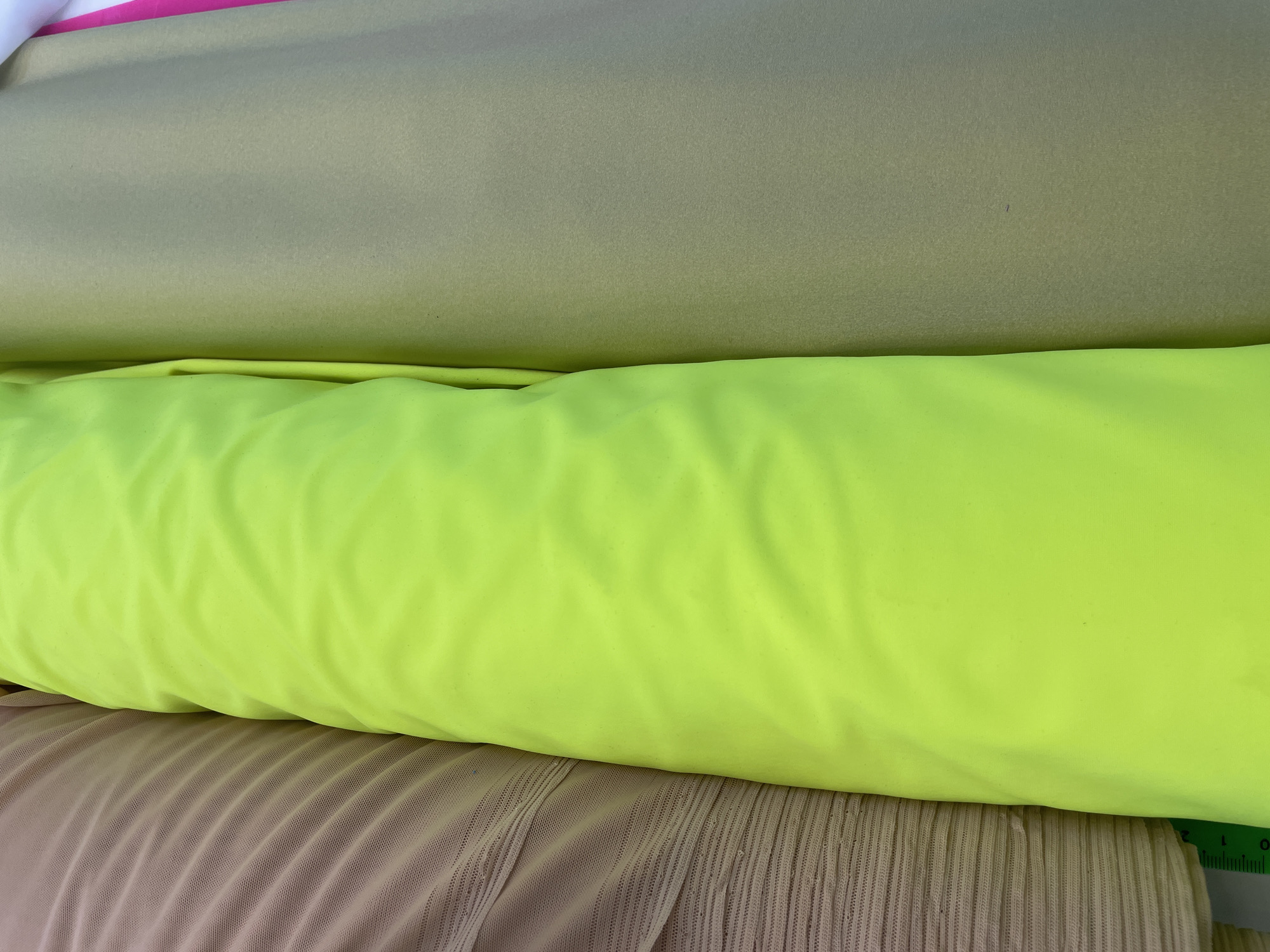 A close-up, eye-level shot displays three large rolls of fabric. The leftmost roll is a pale tan, sheer mesh fabric with fine, horizontal pleats. To its right is a brightly colored roll of neon yellow fabric, tightly rolled and exhibiting soft, undulating folds. The third roll, on the far right, is a dull olive green fabric, also tightly rolled and with a smooth, matte finish. A small portion of a green measuring tape with black markings is visible at the bottom left of the frame. The background is out of focus, suggesting an indoor setting, possibly a fabric store or workshop. There are no people present, and the scene appears static. The location is not determinable from the image content.