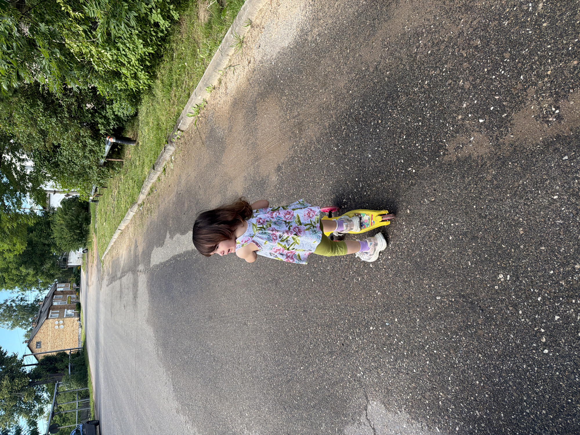 A young child is pictured on a paved surface, possibly a street or a pathway. The child, facing left, is wearing a floral-print dress over green leggings and white sneakers with pink accents. They are positioned on a bright yellow scooter with two wheels visible. The child’s long, brown hair is down, and their expression suggests a moment of play or concentration.
The surrounding environment includes a strip of grass and trees to the left of the paved area, with lush green foliage. Further in the background, a multi-story brick building with several windows is visible, suggesting a residential area. Another, lighter-colored building can be seen behind the trees. A black pipe or drainpipe is also visible in the grassy area. The paved surface appears wet in places, with light reflecting on it, indicating recent rain or damp conditions. The lighting suggests it is daytime, likely late afternoon or early evening given the shadows. The overall scene conveys a sense of casual outdoor activity.