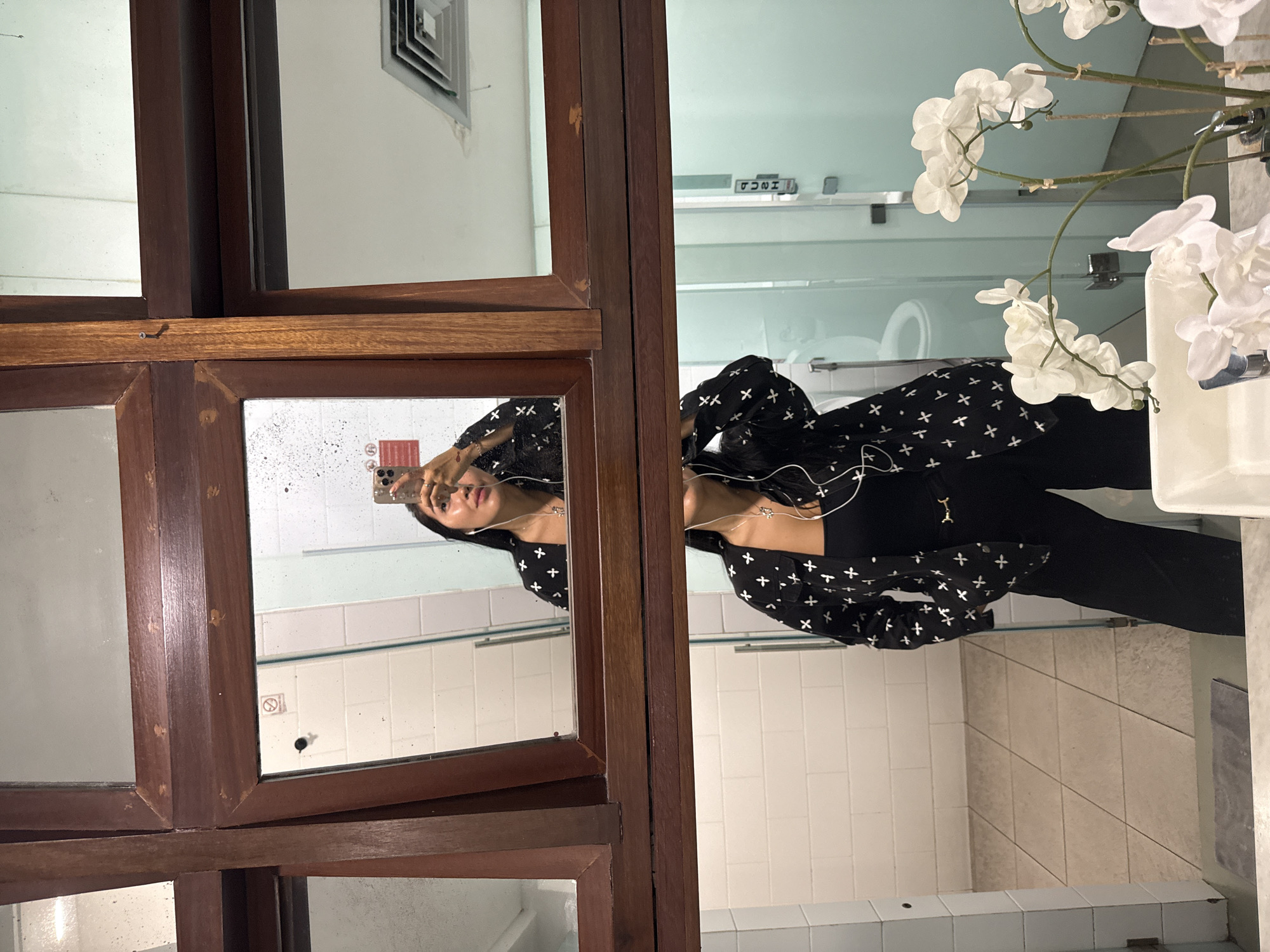 A woman, viewed from the chest up in a mirror reflection, stands in a public restroom. She is taking a selfie with her phone, which is held in her right hand. She is wearing a black blazer with a white cross pattern, a black top, and black pants. She also has white earbuds in her ears. The restroom has white tiled walls and a grey tiled floor. A mirror hangs on the wall, divided into sections by wooden frames. In the foreground, a sink is visible with a vase of white orchids. The woman's reflection is visible in the mirror, as well as a reflection of a toilet and a door with a "PUSH" sign. The location is Kuta, Indonesia.