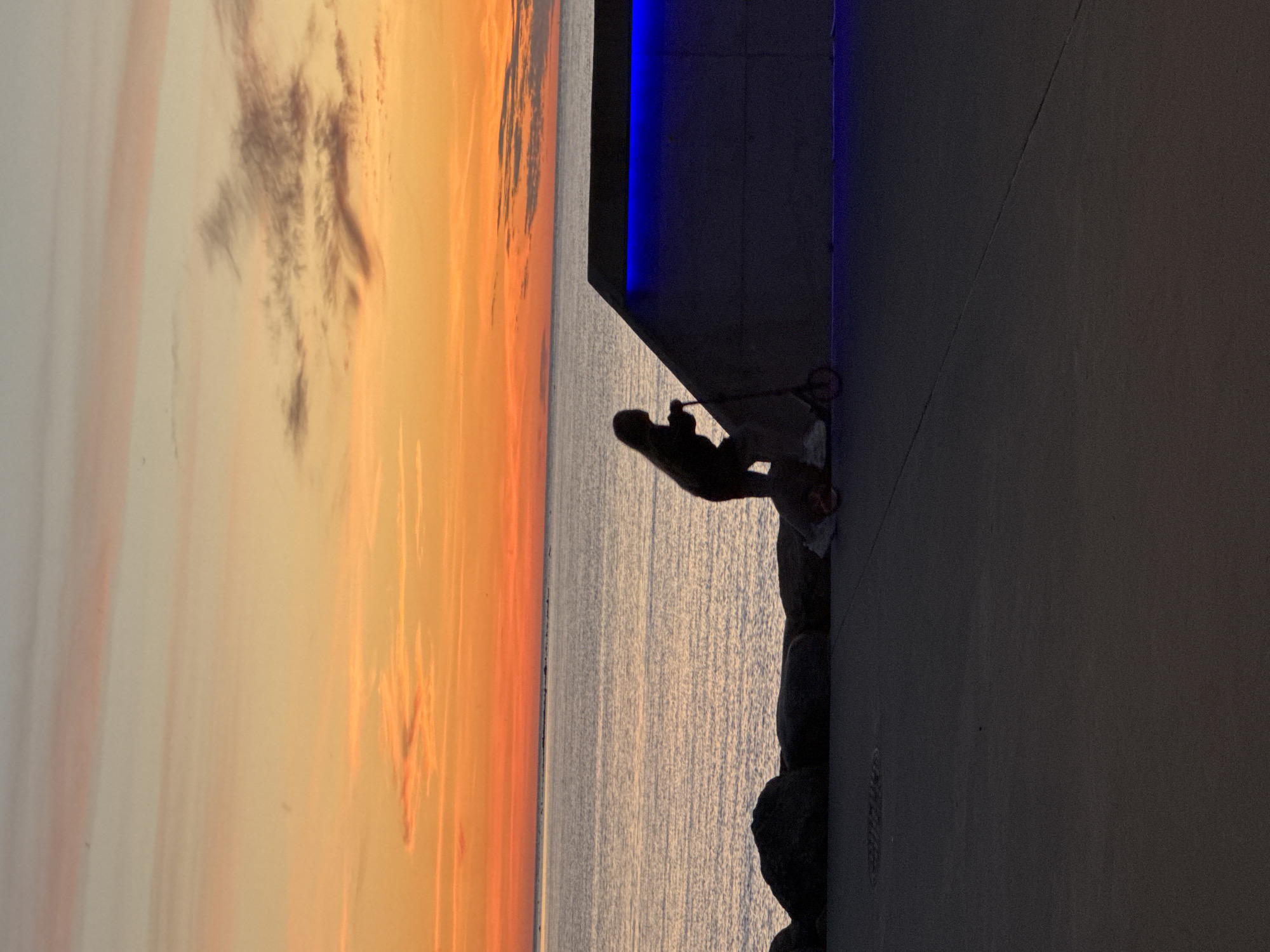 A silhouette of a person riding a scooter is seen against a sunset sky. The sky is a gradient of orange, pink, and pale blue, with wispy clouds. The person is facing left and is in mid-motion on a scooter. To the right of the person is a dark, angular structure illuminated by blue LED lights. In the background, a calm body of water, likely the sea, stretches to the horizon. Large, dark rocks are visible along the edge of the water. The ground in the foreground is paved. This scene appears to be located on a waterfront promenade, possibly in Tallinn, Estonia, given the architectural style of the illuminated structure.