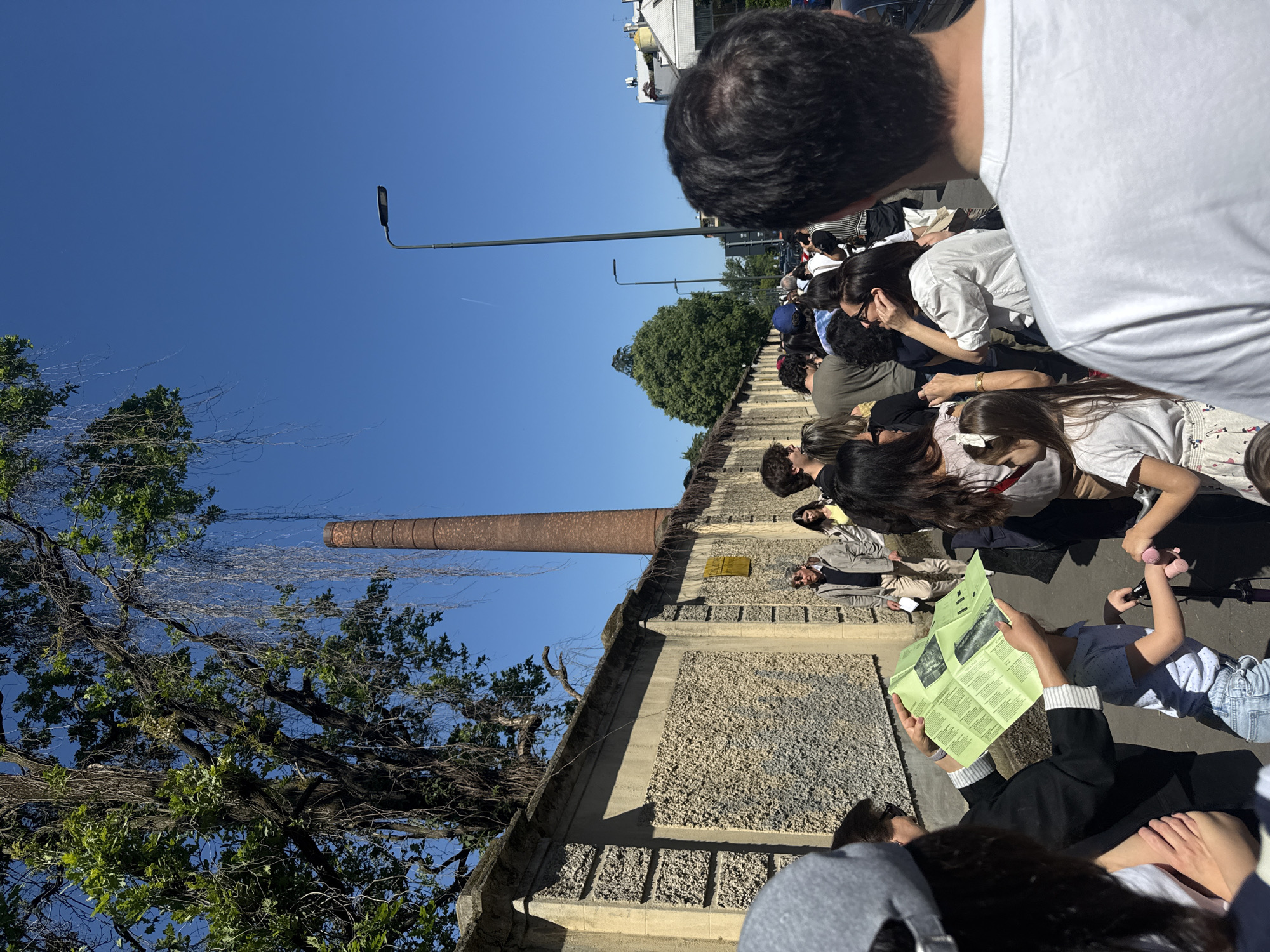A ground-level, angled view of a crowd of people seated along a wall on a sunny day. The wall is made of light-colored stone and runs along the side of a building. A tall, thin, brick smokestack rises from the building behind the wall. A large tree with bare branches extends over the wall and towards the smokestack. In the background are a tree and street lamps against a bright blue sky. The focus is on the seated people, who are looking towards something not visible in the frame. One person is reading a folded green paper or map, while a child nearby holds a pink object. The image shows the group of people in Milan, Italy, gathered to watch an event.