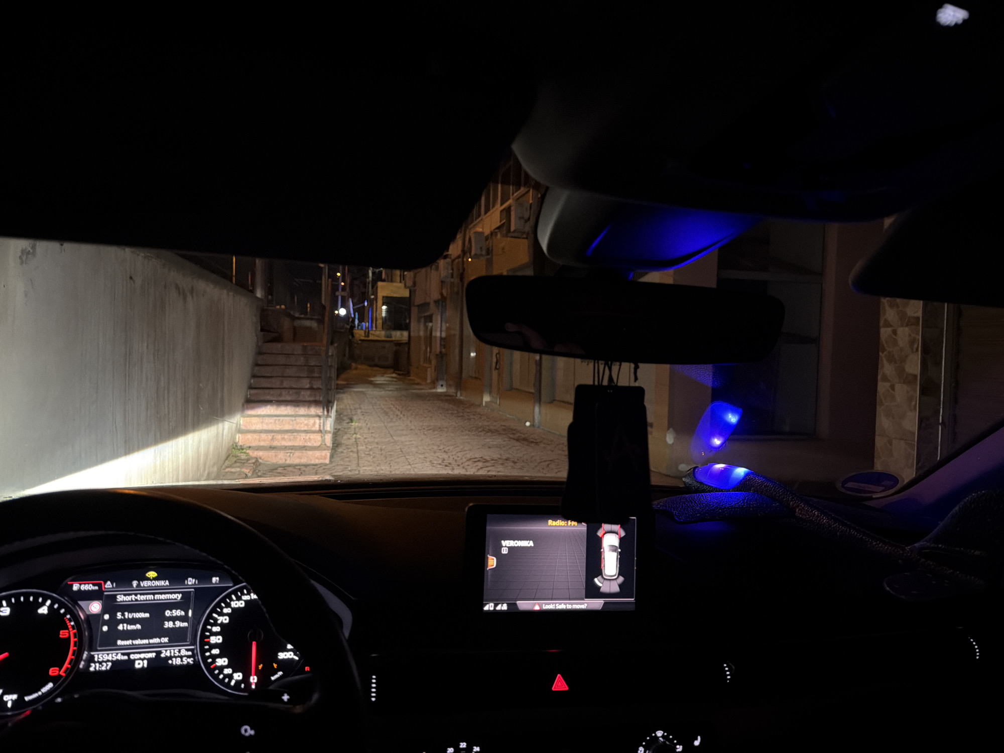 The image shows the interior of a car, presumably an Audi, parked on a narrow street at night. The street appears to be residential, with buildings on both sides. The car's headlights illuminate the street ahead, and there is a set of stairs leading up to a building on the left. The dashboard of the car is visible, displaying information such as speed, fuel consumption, and distance traveled. The car's infotainment system is also visible, showing a parking assist screen. Based on the dashboard data, the car is located in Verona. The time is 21:27, the outside temperature is 18.5 degrees Celcius and the car has travelled 159,454 kilometers.