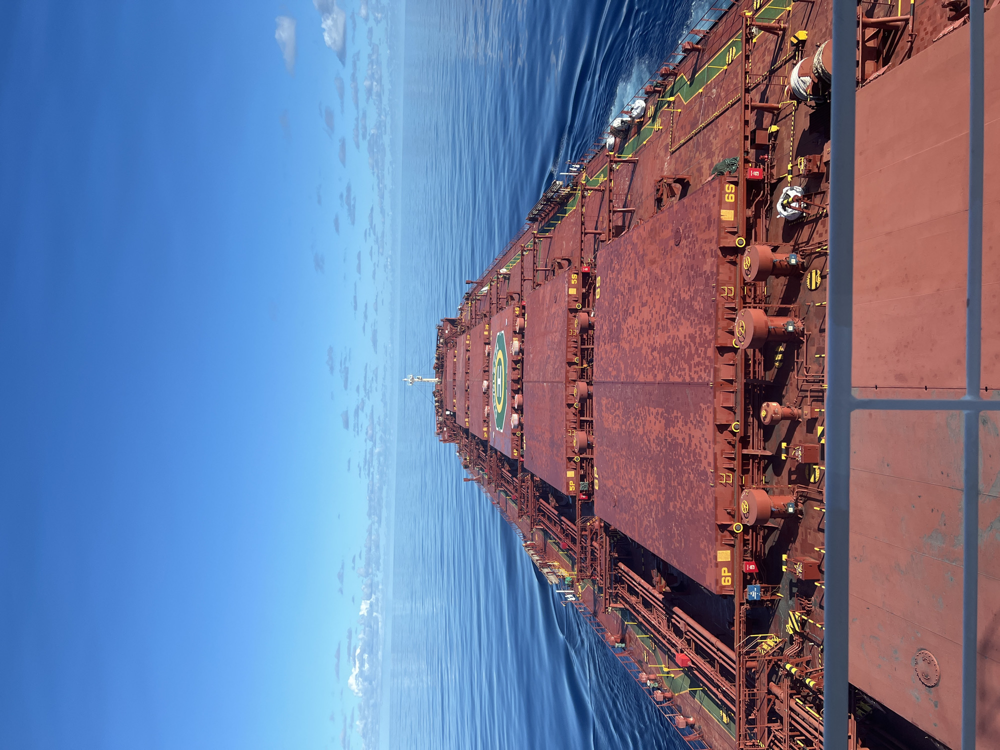 The image depicts a large red cargo ship sailing on a calm, deep blue ocean under a clear sky with scattered white clouds. The perspective is from the upper deck of the ship, looking forward along its length. The ship's deck is covered in numerous hatch covers, pipes, and mechanical equipment. Markings such as "6P" and "6S" are visible on some of the hatch covers. The water surrounding the ship shows a visible wake. The horizon line is sharp, separating the expansive ocean from the sky. The scene suggests a vessel in transit at sea, with no urban or city elements present.