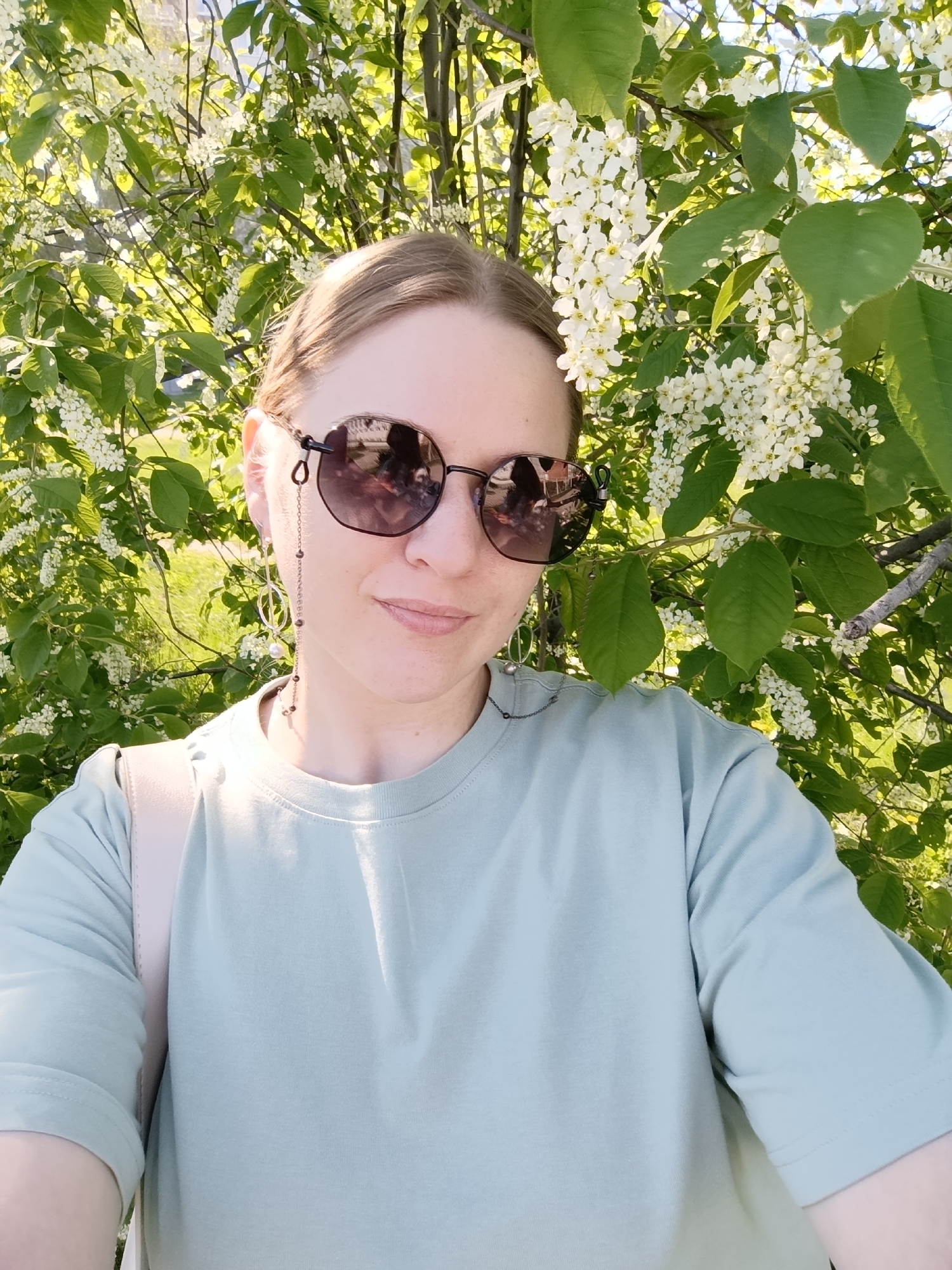 The image shows a selfie of a woman wearing sunglasses and a light green t-shirt. She is positioned in front of a tree laden with white blossoms and green leaves. A shoulder strap of a bag is visible on her left shoulder. The location appears to be outdoors, possibly in a park or garden, but the city and specific location are unknown.