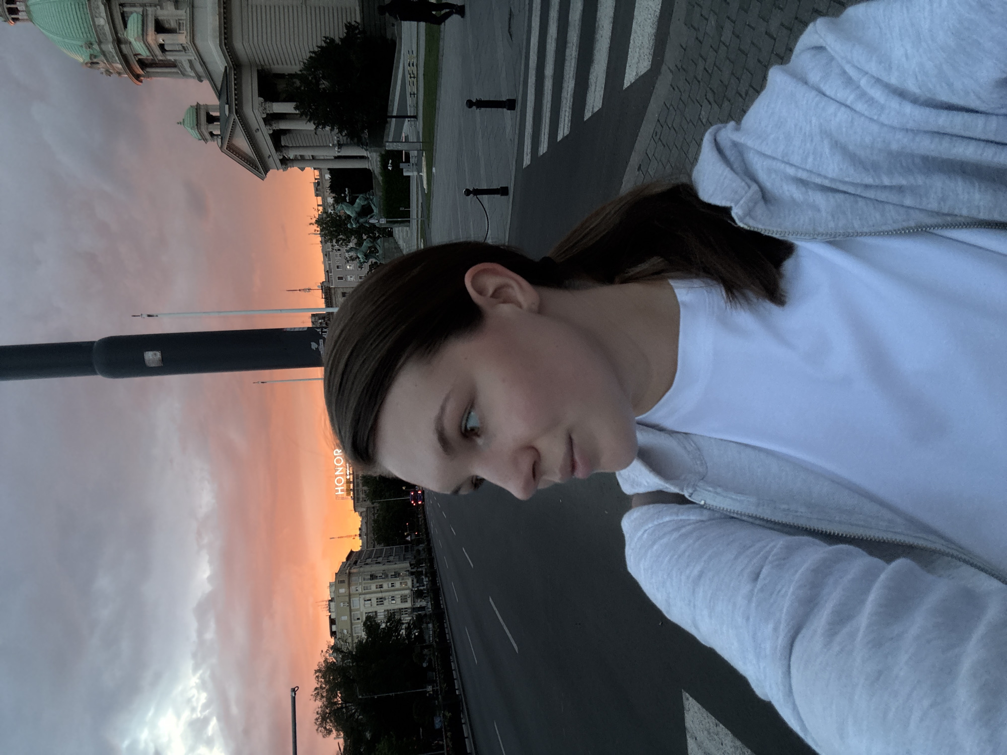 A medium shot, selfie-style, captures a young woman from the chest up, looking to her left. She has fair skin, brown hair pulled into a ponytail, and is wearing a light grey hooded jacket over a white t-shirt. The background depicts a street scene at dusk in Vračar, Belgrade, Serbia. A multi-lane road with white lane markings dominates the left side of the frame, with cars visible in the distance. Buildings line the street, and a prominent "HONOR" sign is illuminated in the distance. To the right, a grand, classical building with columns and a green dome, likely the National Assembly of Serbia, is partially visible. A pedestrian crossing with white stripes is in the foreground, along with bollards and a chain. The sky is dramatic, filled with orange and grey clouds, indicating sunset. The overall lighting is dim, typical of twilight.