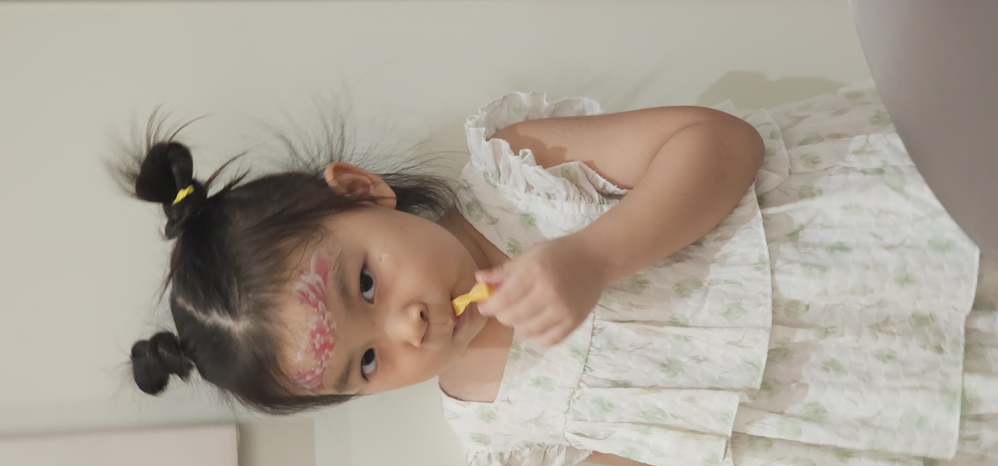 A young East Asian girl with dark hair styled into two messy buns and face paint on her forehead is holding a yellow object to her mouth as if brushing her teeth. She is wearing a white ruffled dress with a green floral pattern. The background is a plain, light-colored wall. The image appears to be taken indoors.