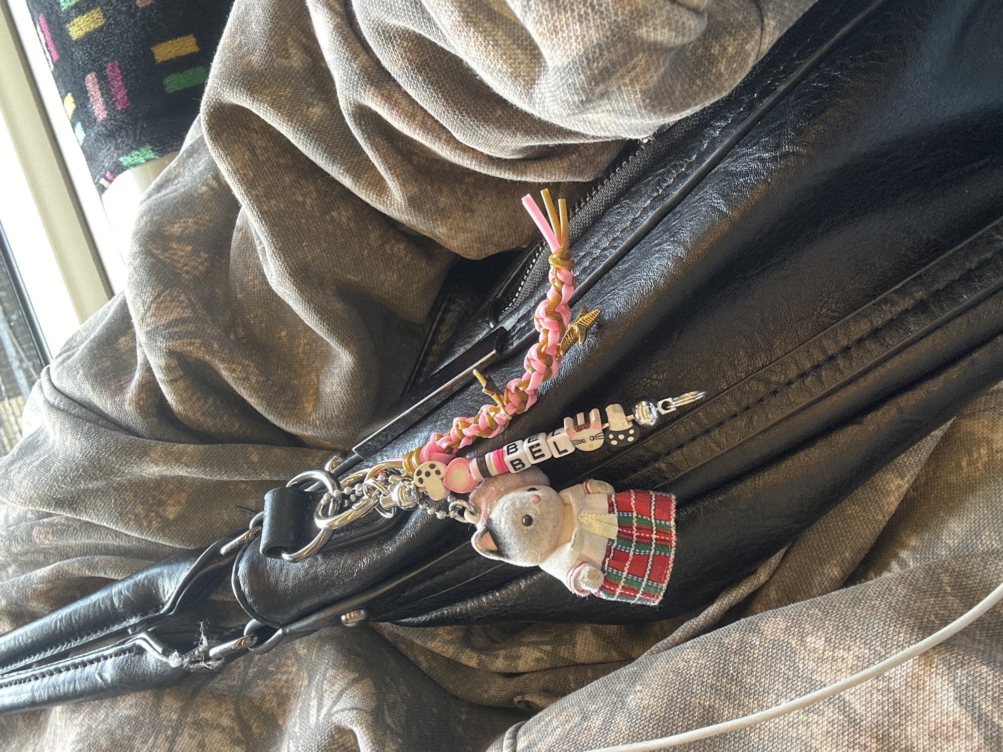 The image shows a close-up of a black leather bag or jacket lying on what appears to be a patterned seat or fabric surface, possibly in a public transport setting. The bag has several keychains attached to it, including a pink braided keychain, a keychain with letter beads spelling "BEL," and a plush toy in the shape of a gray mouse wearing a white and red plaid dress. The overall setting suggests a casual or travel scene, with a focus on personal belongings. The location in London, United Kingdom, cannot be definitively determined from the given image.