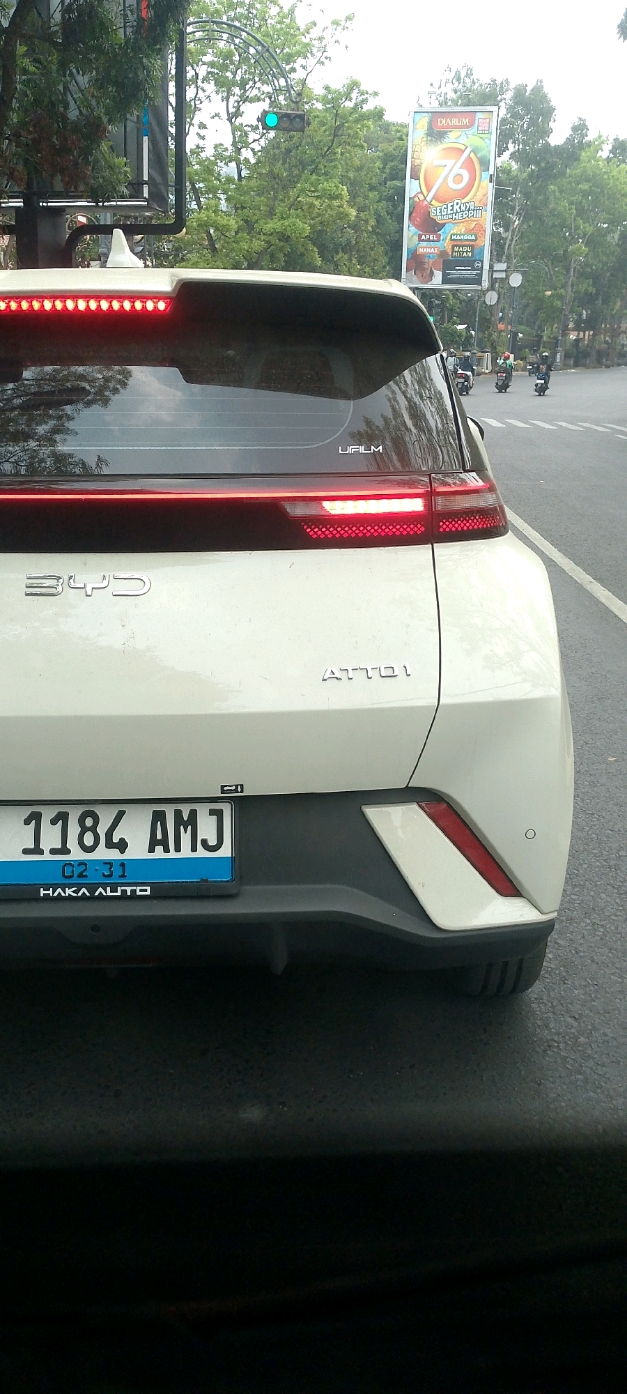 A rear view of a white BYD ATTO 1 electric car is captured from a slightly low angle, showing the car from the bumper up to the rear spoiler, which is illuminated with red LED lights. The car's license plate, "1184 AMJ", is clearly visible below the rear lights. The BYD logo is present on the left side of the rear, and the "ATTO 1" model designation is on the right. The car is on a paved road with other vehicles and a traffic light in the background. A billboard for "Djarum" (a tobacco company) featuring a number "76" and various fruits is visible on the right side of the image. The scene appears to be in an urban environment with trees and buildings lining the street, indicative of a location within Bandung City, Indonesia, likely a relatively busy street with traffic. The overall lighting suggests it is daytime.