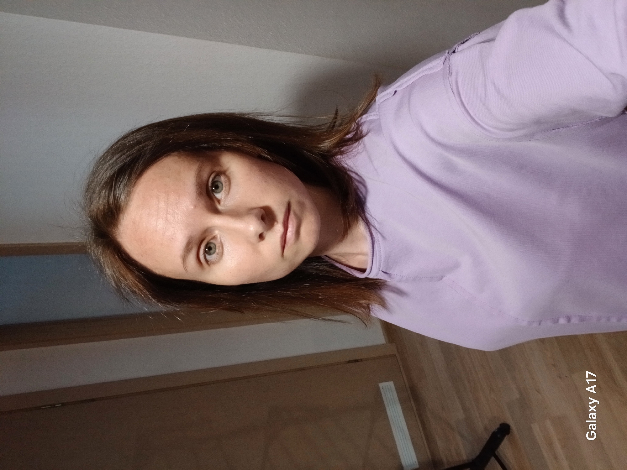 A woman with brown hair and light eyes is facing the camera. She is wearing a light purple shirt. The background shows a partially visible wooden door and a light-colored wall. The floor is made of wooden planks. The text "Galaxy A17" is present in the bottom right corner of the image. There is no information to suggest this image was taken in Yekaterinburg, Russia.