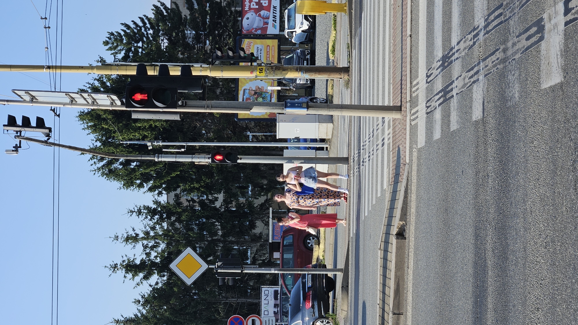 The image captures a street scene in Prešov, Slovakia, focusing on a pedestrian crossing. Three young women stand on the sidewalk awaiting a signal to cross. The woman on the left wears a striped red and white dress and holds her arm to her face. The middle woman is in a floral print dress, and the woman on the right is wearing a white tank top and denim shorts. A traffic light above them displays a red pedestrian signal, indicated by a red silhouette of a person. The background shows a row of tall coniferous trees, traffic lights, utility poles, and buildings. Cars are visible on the street, including a black Mercedes-Benz sedan and a red minivan. The asphalt road features a pedestrian crossing with white stripes. The sky is clear and blue, suggesting a sunny day.