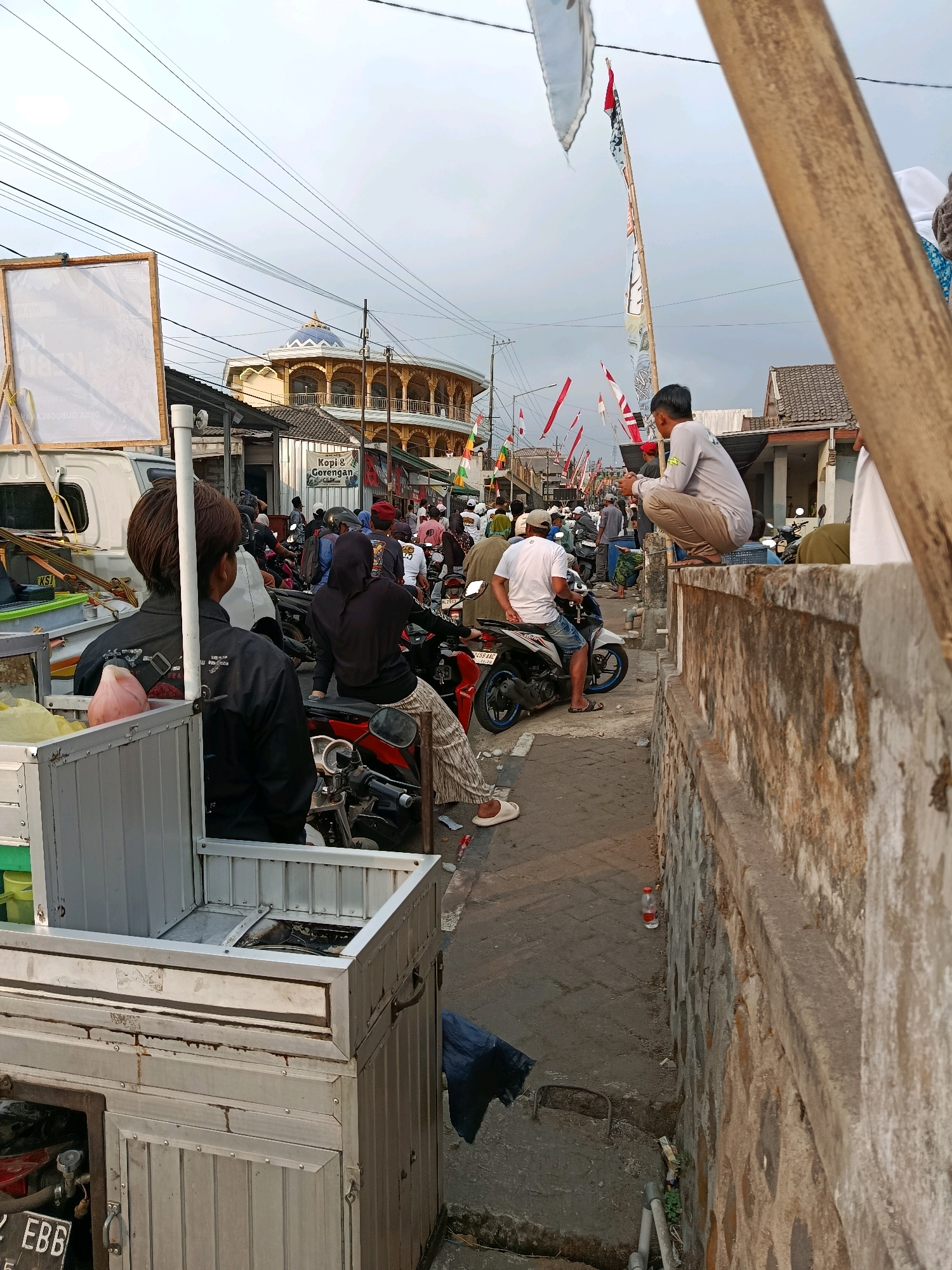 The image depicts a street scene in Gubukklakah, Indonesia, likely during a festive occasion, given the presence of numerous flags and banners. The street is lined with buildings and crowded with people and motorcycles. In the foreground, a metal food cart is partially visible on the left. A person in a black jacket is standing behind the cart, facing away from the camera. Further down the street, several people are gathered, some on motorcycles, and others standing or walking.
A prominent circular building with an ornate dome and arched windows is visible in the background. Flags and pennants are strung across the street, fluttering in the breeze. On the right side of the image, a stone wall runs along the street, with a person squatting on top of it, looking towards the street. The overall atmosphere suggests a busy public gathering, possibly a market or a celebration. The sky is overcast, indicating potentially inclement weather.