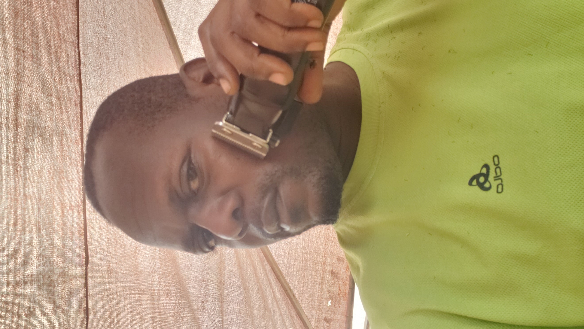 A man is shaving his face with an electric trimmer in Narayi, Nigeria. He is wearing a light green t-shirt with a logo. The background is a light brown fabric that appears to be a covering. The man is looking at the camera. There are small clippings of hair on his t-shirt.