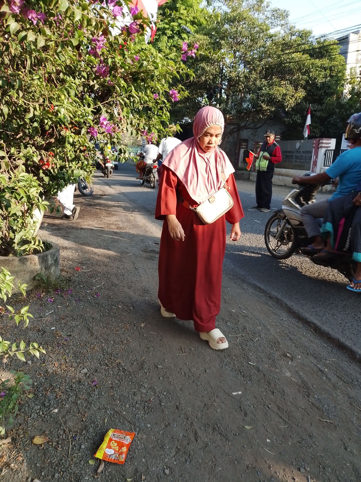 In Pasuruan, Indonesia, a woman in a maroon abaya and pink hijab walks along a gravel path next to a paved road. She carries a beige crossbody bag. A red and white flag is visible in the background, as are several trees and buildings. A man in a red jacket and a reflective vest stands nearby, also holding a small flag. Several motorcycles are parked or moving on the road, and one person rides on the back of a motorcycle. A crumpled snack wrapper lies on the ground in the foreground. The scene appears to be in an urban or suburban area during daylight hours.