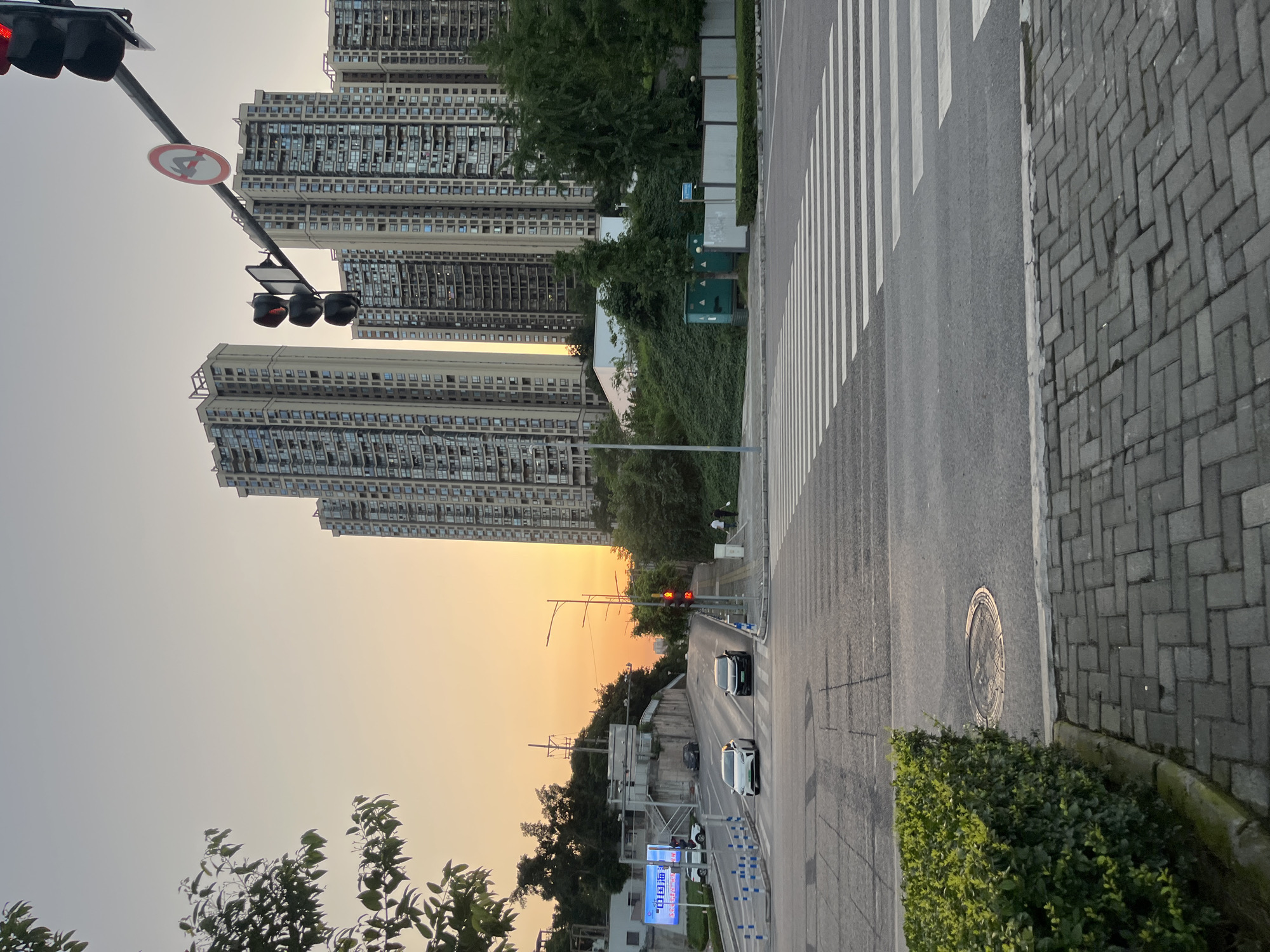 The image shows a street scene in Banan, China, at sunset. The street has a crosswalk and is lined with tall apartment buildings. There are cars driving on the street, and a traffic light is visible. The sidewalk is paved with grey rectangular pavers. A shrub lines the road.