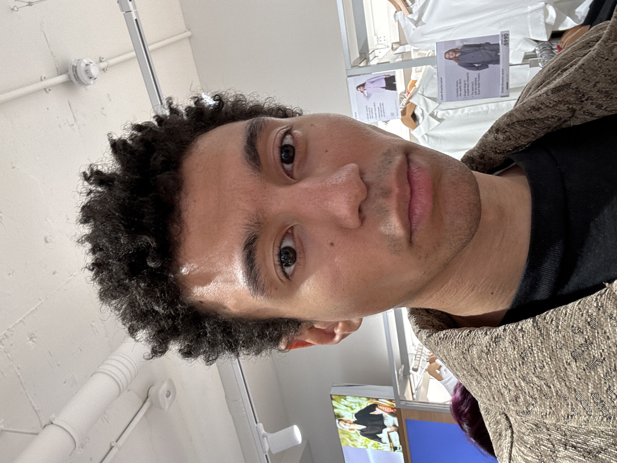 A selfie shows a light-skinned man with tightly curled, dark hair and brown eyes. He is wearing a black t-shirt and a brown patterned jacket. The background appears to be inside a store, possibly a clothing shop. Racks of white shirts and clothing are visible, along with promotional signs displaying images of people wearing garments and their prices. The location is not discernible as being in London, United Kingdom, without additional visual cues.
