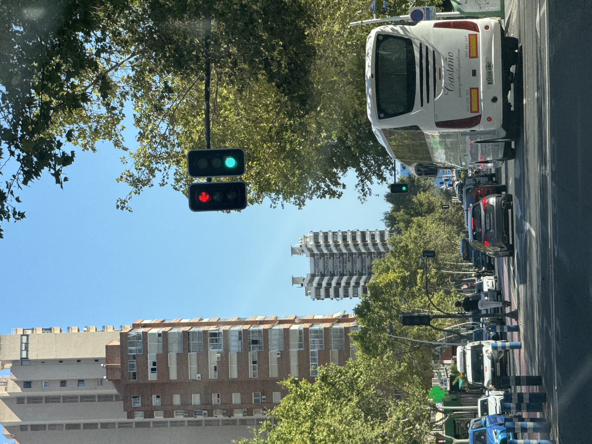 This is a street scene in Benidorm, Spain, captured from a vehicle moving forward. The image features a bright, sunny day with a clear blue sky. In the foreground, a white tour bus with "Castaño" written on its rear is prominent, traveling in the right lane. Several cars are visible in front of and behind the bus, as well as in other lanes. A person is riding a scooter on the road in the middle distance.
On the left side of the street, tall residential buildings rise high into the sky, characterized by their beige and brick facades, balconies, and numerous windows. Some of these buildings appear modern, while others have a more classic architectural style. Green trees with dense foliage line the street, providing shade and contributing to a lush urban environment.
Traffic lights are visible above the road, with one showing a red light and another a green light. These indicate the flow of traffic. The road itself is paved and marked with white lines, suggesting a well-maintained urban infrastructure. The overall impression is of a bustling city street with active traffic and a blend of modern and older architecture, characteristic of a tourist destination like Benidorm.