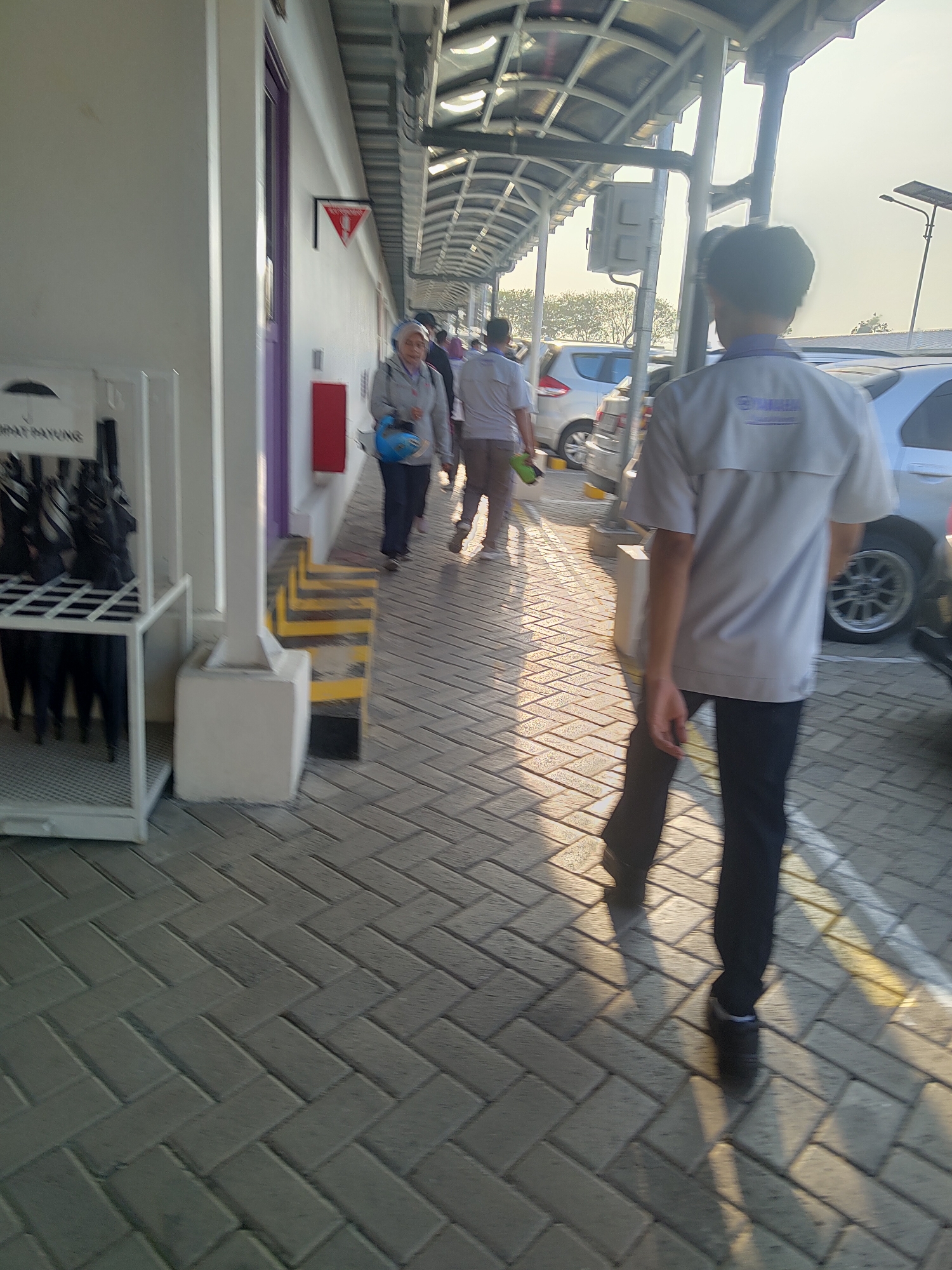 A group of people are walking along a covered walkway with a tiled floor in Raci, Indonesia. A person in a light-colored shirt and dark pants is walking away from the camera. To their left, a woman wearing a headscarf and a blue helmet is walking. In the background, cars are parked. There is an umbrella stand on the left side of the image. The sun is shining, casting long shadows on the ground.