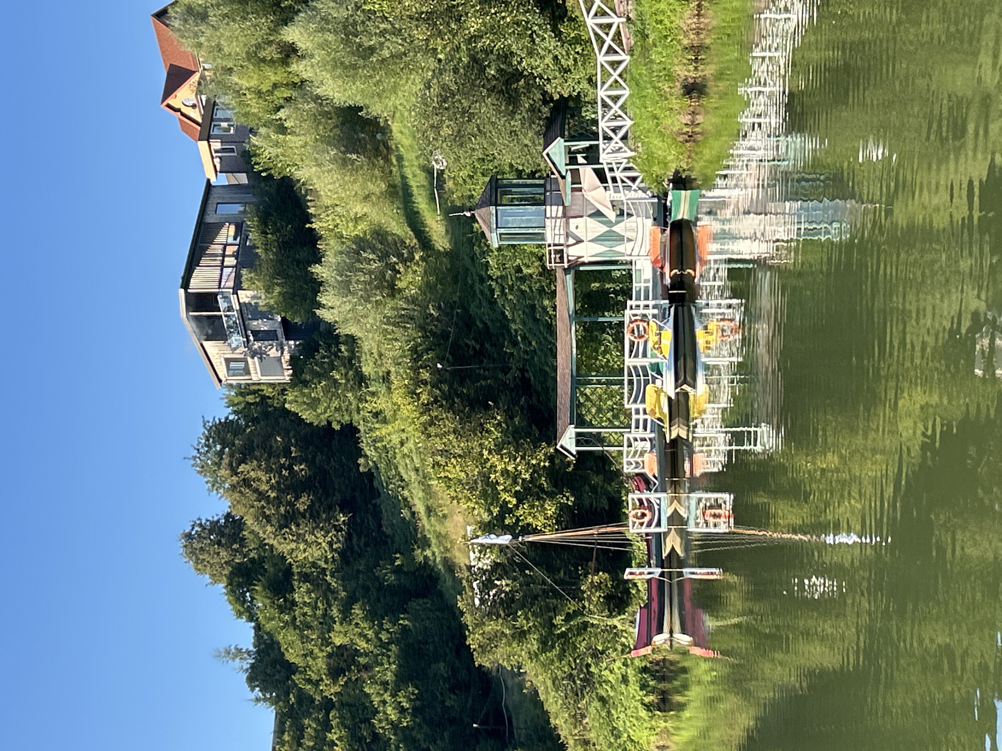 The image depicts a serene waterside scene, likely within the city of Romashkovo, Russia. The foreground is dominated by a tranquil body of water, reflecting the lush greenery and structures on its bank. Several small boats, including pedal boats and what appears to be a traditional wooden vessel with a mast, are docked at a wooden pier. The pier itself is adorned with a white latticed fence and a structure resembling a small lighthouse or observation tower with a green roof. Behind the pier, the landscape rises into a verdant hill densely covered with trees. Perched atop the hill are two modern houses, one with a dark facade and balconies, and another with a red tiled roof. The sky above is a clear, bright blue, suggesting a pleasant day. There are no people visible in the image.