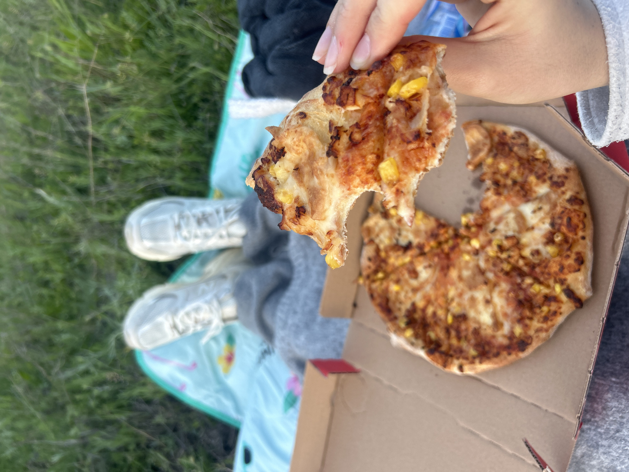 A first-person perspective image shows a woman holding a piece of pizza in her right hand, with the rest of the pizza in a cardboard box to her right. The pizza appears to be topped with corn and other browned ingredients. The woman is wearing a white sweatshirt. In the background, her legs are visible with white sneakers. She is sitting on a light blue blanket with a floral pattern. The ground is covered in green grass. The location is unknown.