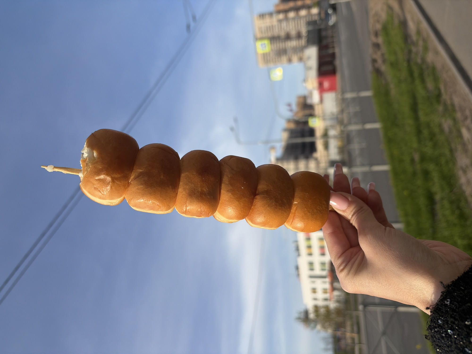 A person is holding a skewer of what appears to be six small, round doughnuts in their right hand. The doughnuts have a shiny, light brown surface. The skewer is visible at each end of the doughnuts. The person has manicured nails. They are standing outdoors with a cityscape in the background. The background is slightly blurred but includes buildings, trees, street lights, and a clear blue sky. The location is Saint Petersburg, Russia, identified by the distinct architecture. The photo is taken during the day, and the natural lighting suggests an outdoor setting.
