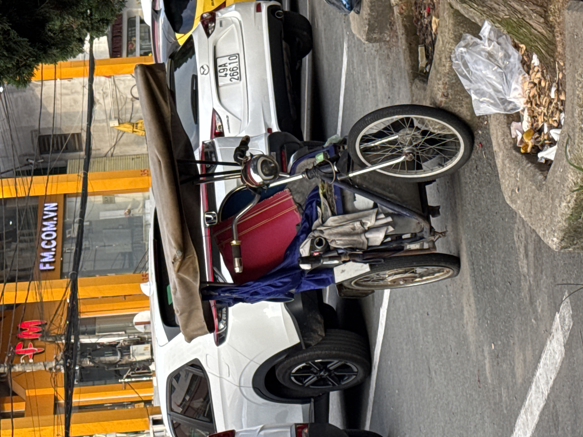 The image shows a street scene in Da Lat, Vietnam, with a three-wheeled motorized vehicle, likely a cyclo or pedicab, parked in the foreground. The vehicle has a covered passenger area with a red seat and a brown canopy. It is laden with blue fabric and gray cloth. Behind the vehicle are several cars, including a white Honda CR-V and a white Mazda with a license plate reading "49A 266.10". In the background, a building with orange accents and signs, including one that says "FM.COM.VN", is visible. The street is paved and bordered by a concrete curb. Litter, including a plastic bag and food scraps, is present on the curb. The overall impression is of a typical urban street with parked vehicles and commercial buildings.